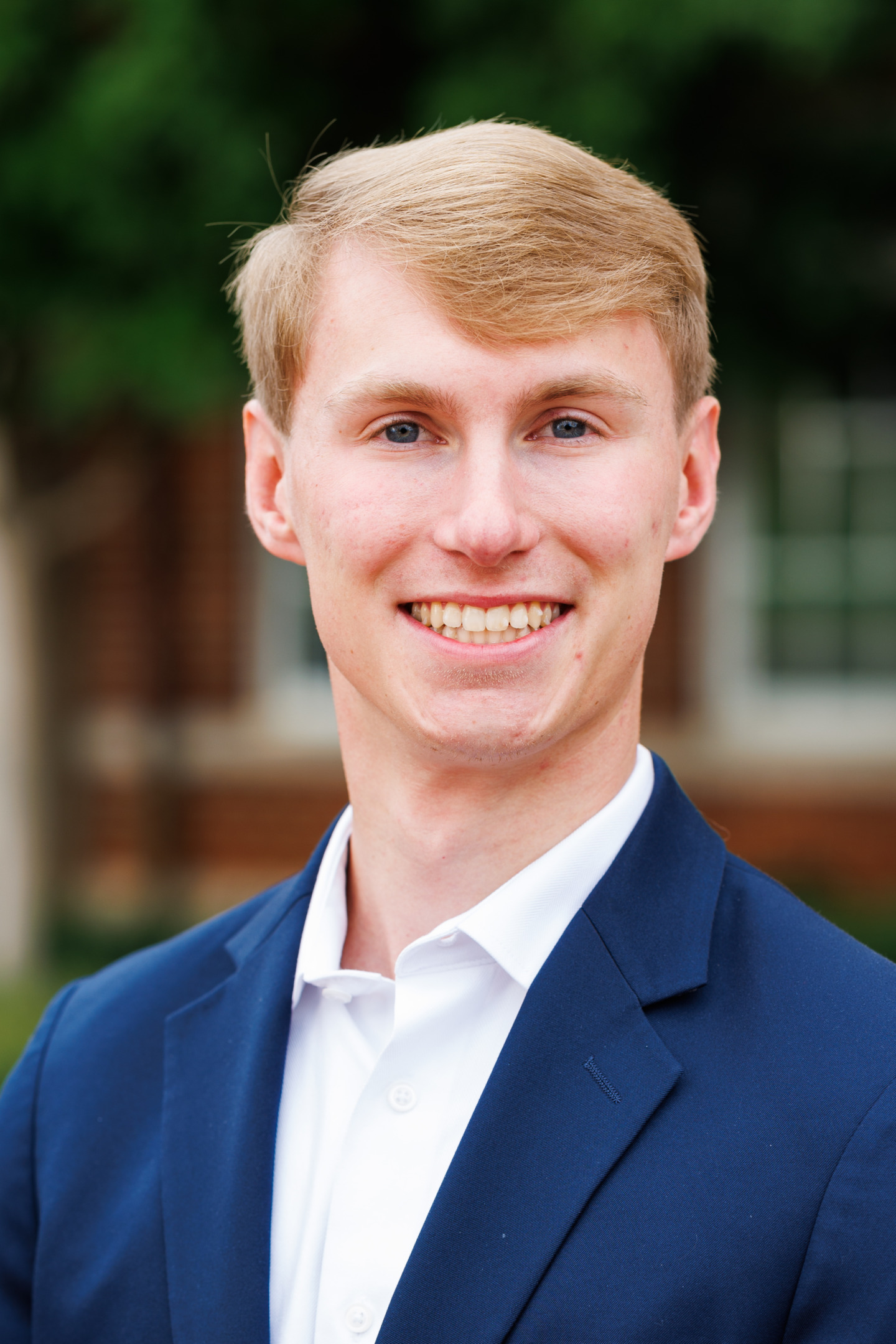 A young person with light hair and blue eyes smiles warmly while wearing a navy blazer over a white collared shirt, set against a blurred outdoor background.