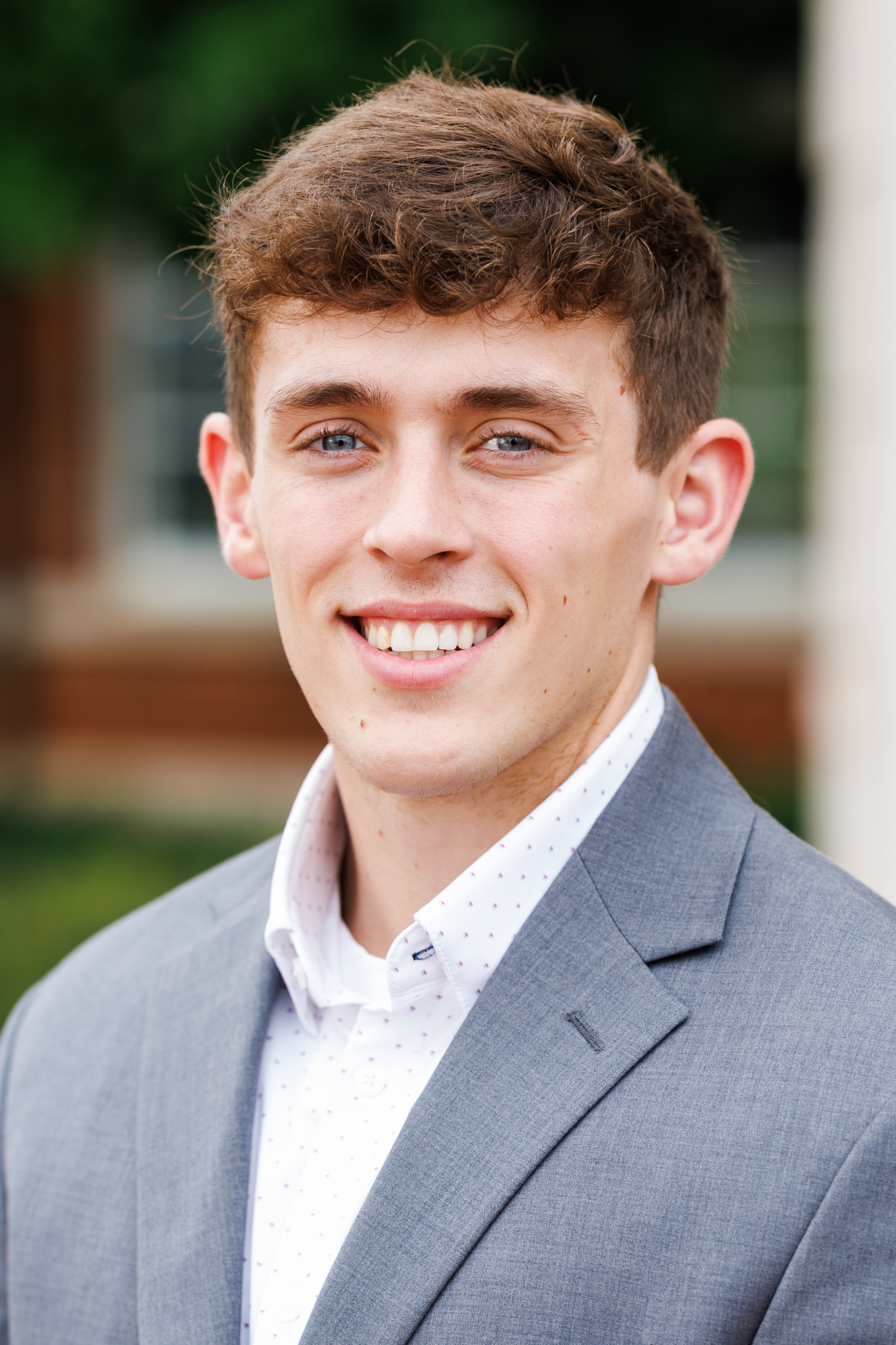 A young person with curly brown hair and blue eyes smiles warmly while wearing a gray suit jacket over a white patterned shirt.