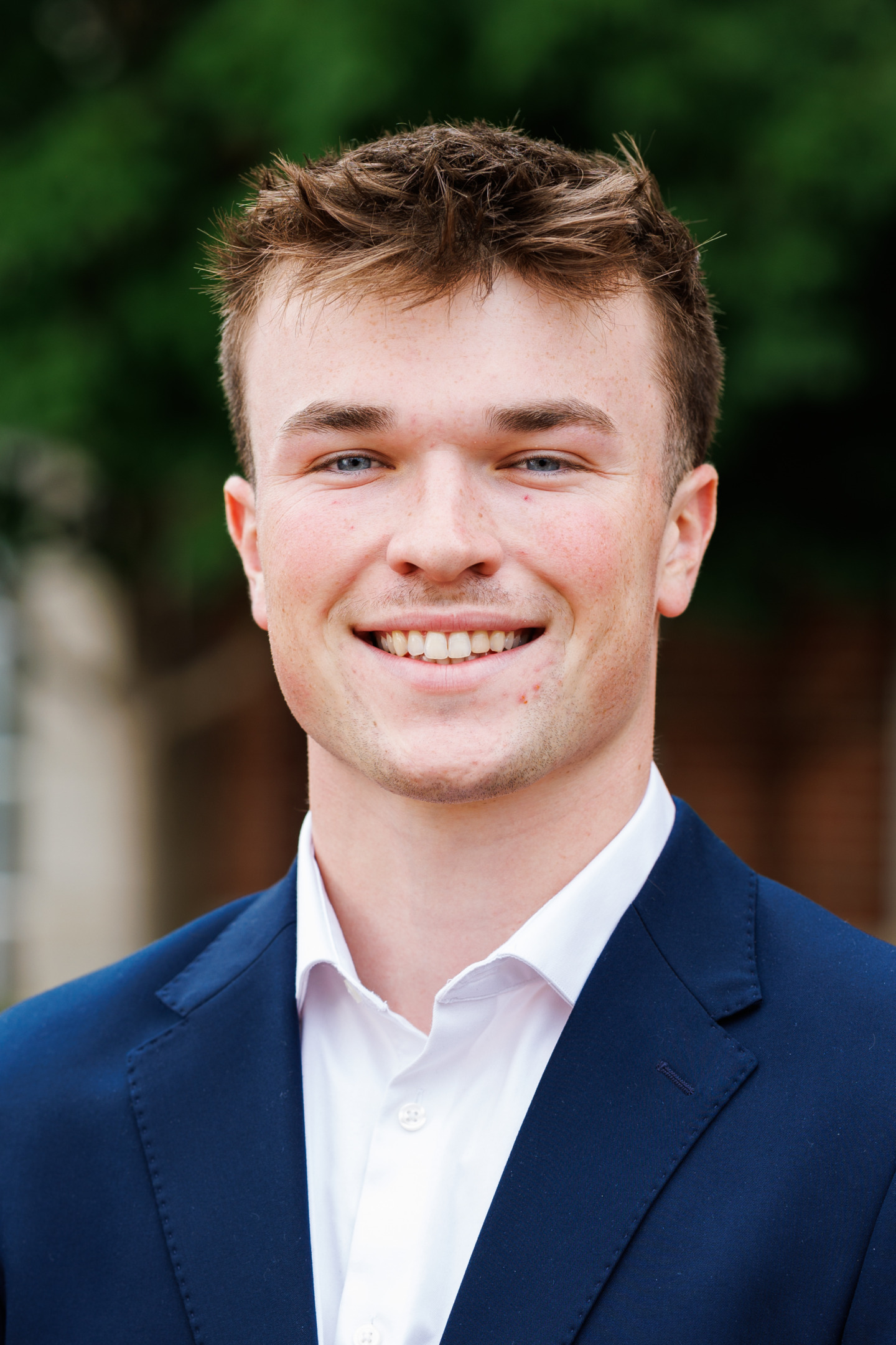 A young person with short brown hair and blue eyes smiles warmly while wearing a navy blue suit jacket over a white collared shirt, set against a blurred green background.