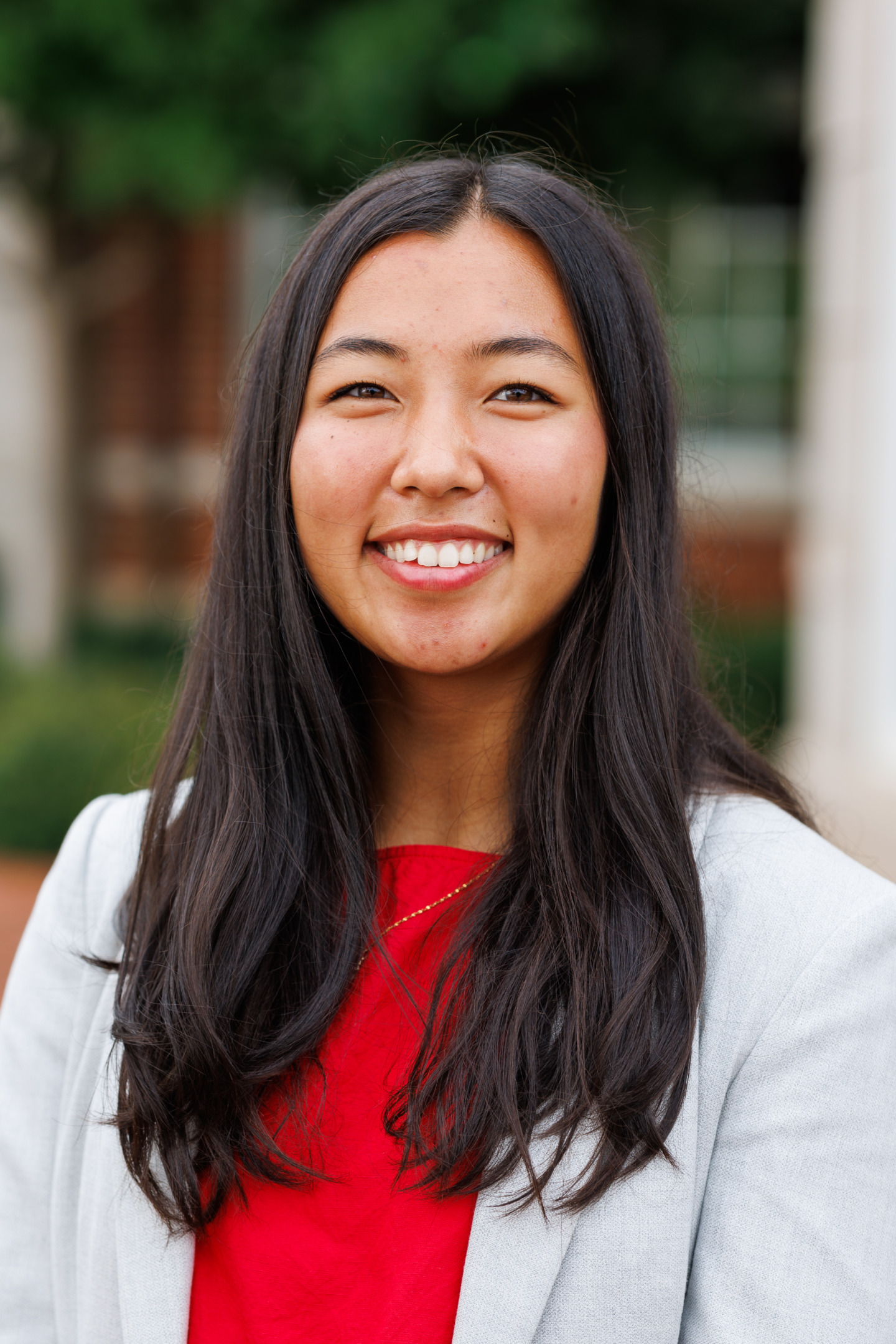 A person with long dark hair smiles warmly while wearing a red top and a light gray blazer, set against a softly blurred outdoor background.