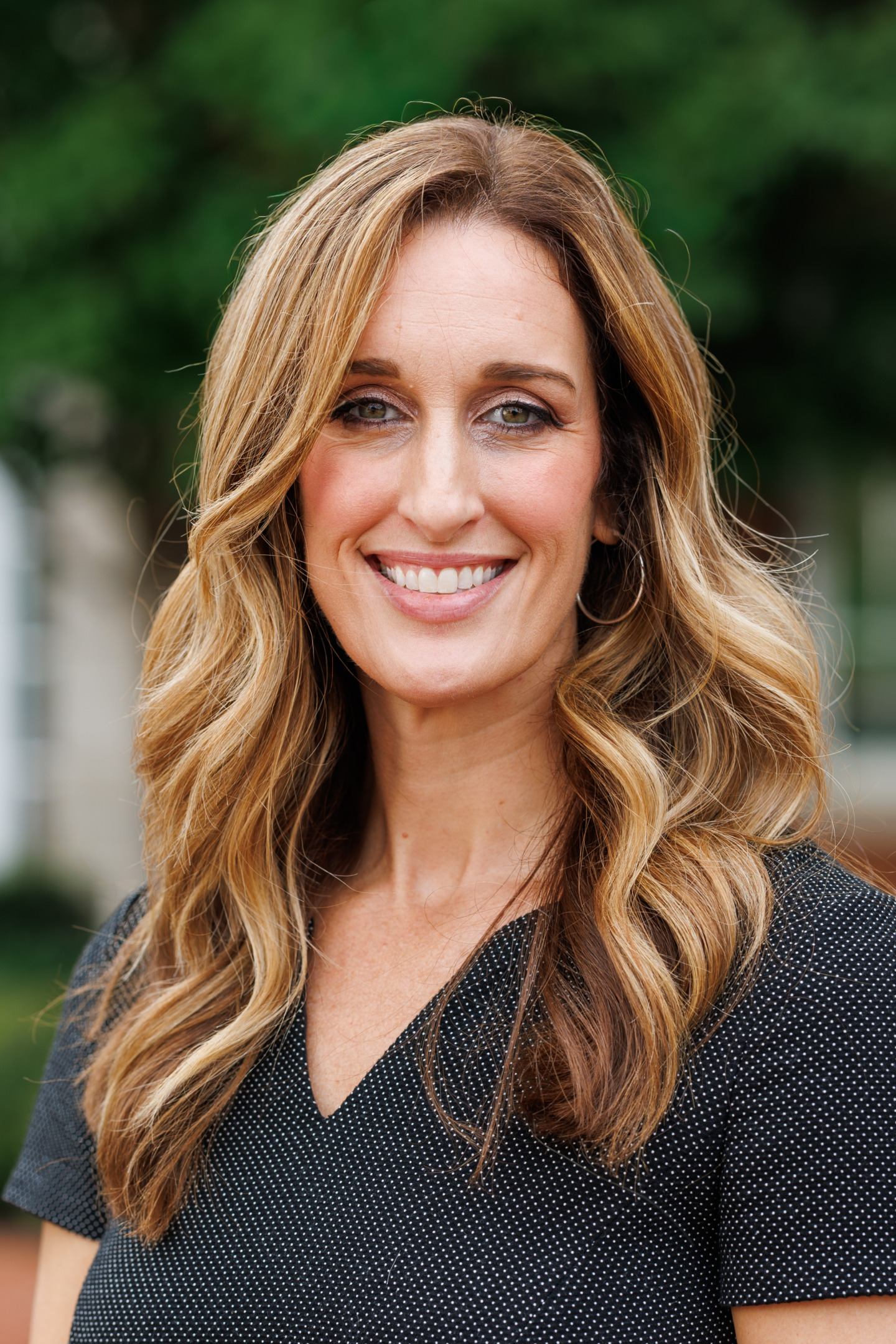 A smiling person with long, wavy blonde hair wearing a black polka-dot top, standing outdoors with greenery in the background.