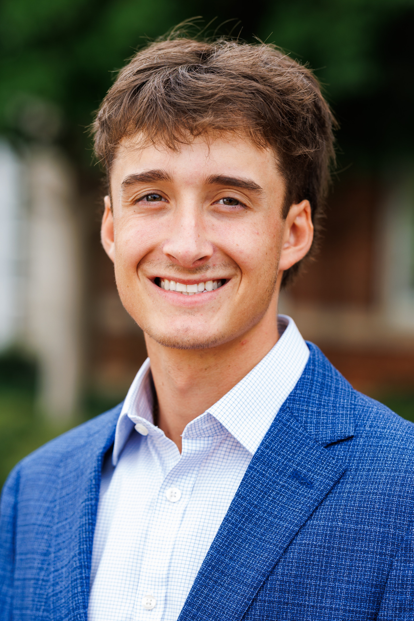 A young person with brown hair smiles warmly while wearing a blue blazer and a light-colored checkered shirt, set against a softly blurred outdoor background.