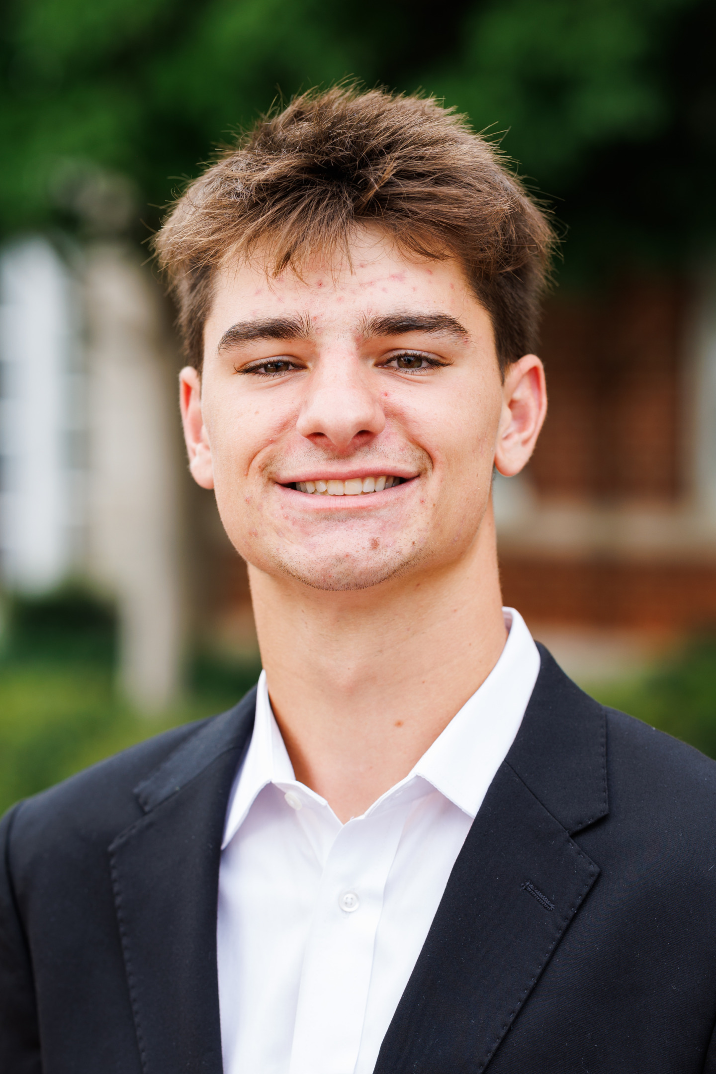 A young person with short brown hair, wearing a dark suit jacket and white shirt, smiles at the camera outdoors.