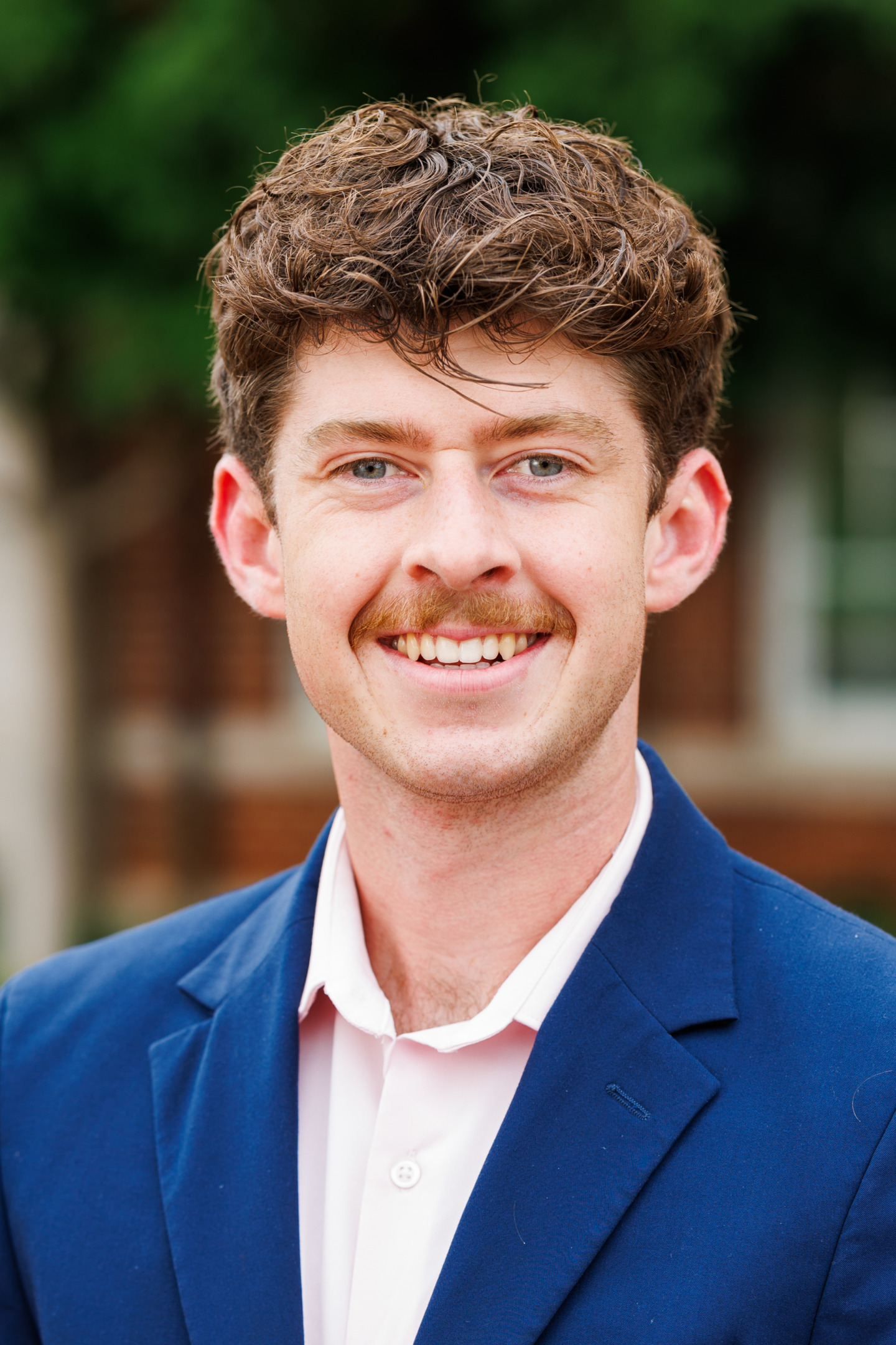A person with curly brown hair and a mustache smiles warmly while wearing a blue blazer over a light pink shirt, set against a blurred outdoor background.