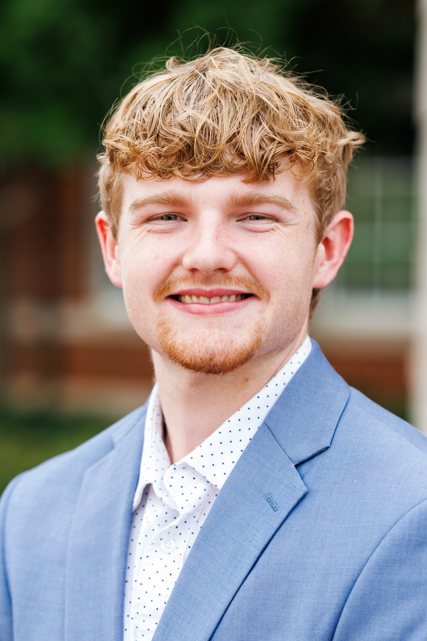 A young person with curly blonde hair and a reddish beard smiles warmly while wearing a light blue suit jacket over a white polka-dot shirt.