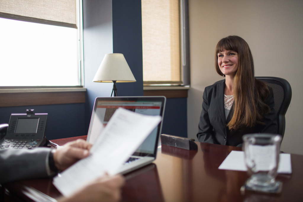 A professional in a suit sits at a desk in an office, smiling while reviewing documents during a meeting.
