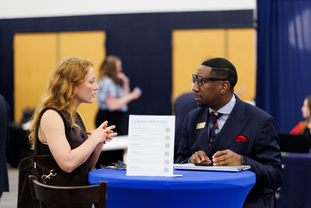 A person in a suit sits at a blue table with a career services sign, conversing with a young individual who is gesturing while speaking.