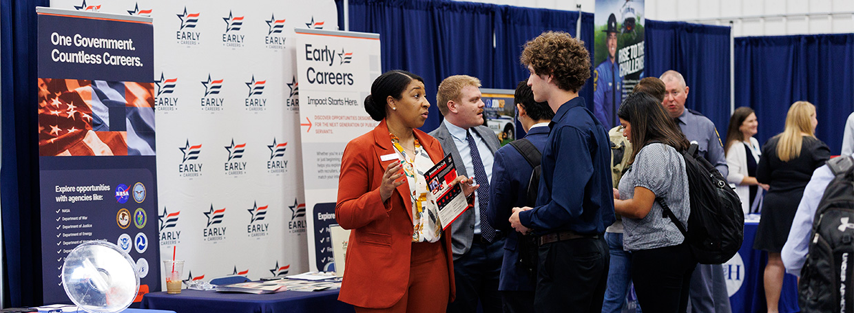 A professional engages with attendees at a career fair booth promoting government job opportunities.