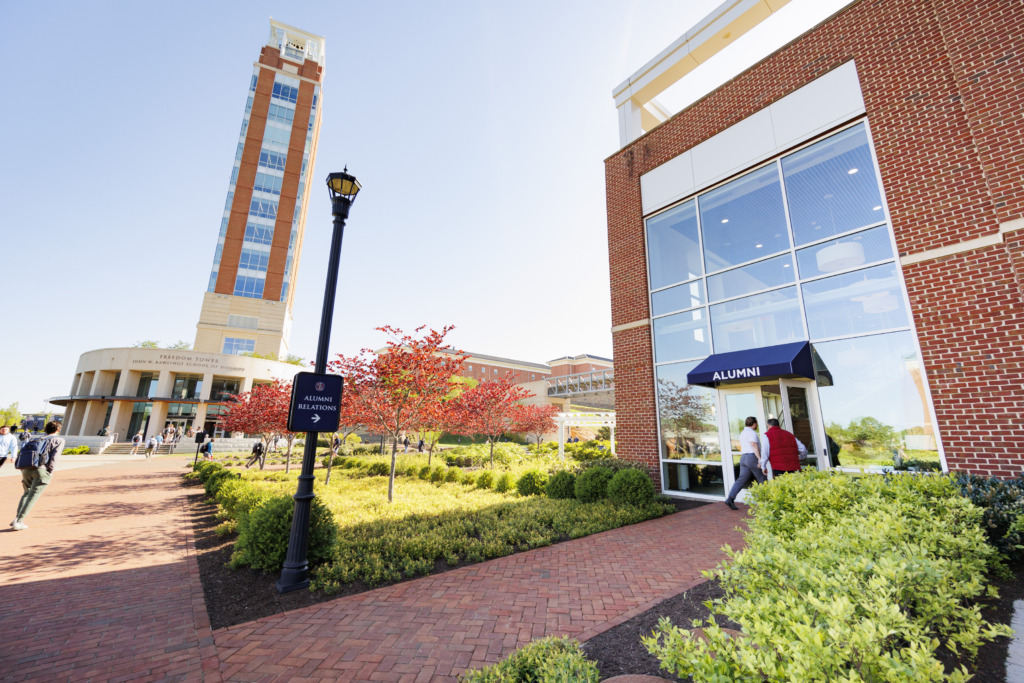 A sunny campus scene featuring a tall tower, brick buildings with glass windows, and people walking along a red-brick pathway lined with greenery and autumn-colored trees.