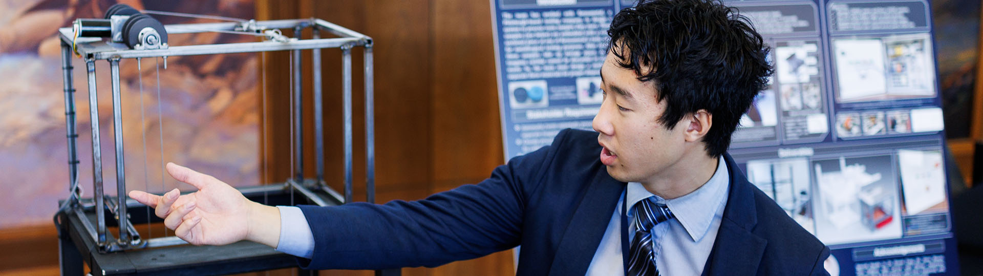 A person in a suit gestures toward a mechanical device on display at a conference, with a presentation board in the background.