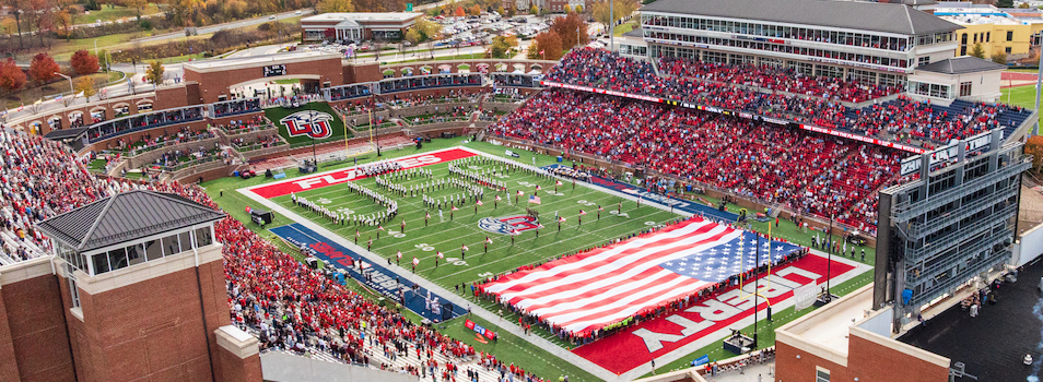 An aerial view of a football stadium filled with spectators, featuring a large American flag and the word "LIBERTY" displayed on the field during a ceremony.