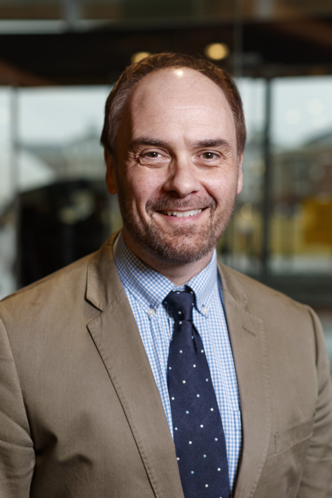 A smiling person in a tan suit, blue checkered shirt, and dark tie with white polka dots stands indoors with a blurred background.