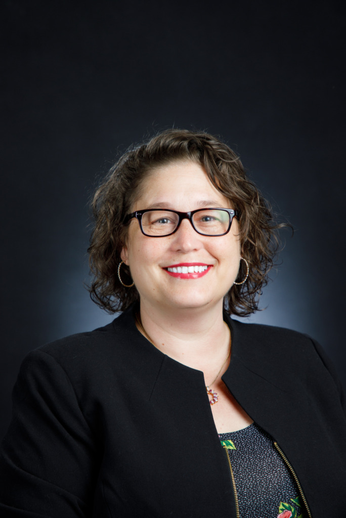A smiling person with short curly hair, wearing glasses and a black blazer, stands against a dark background.