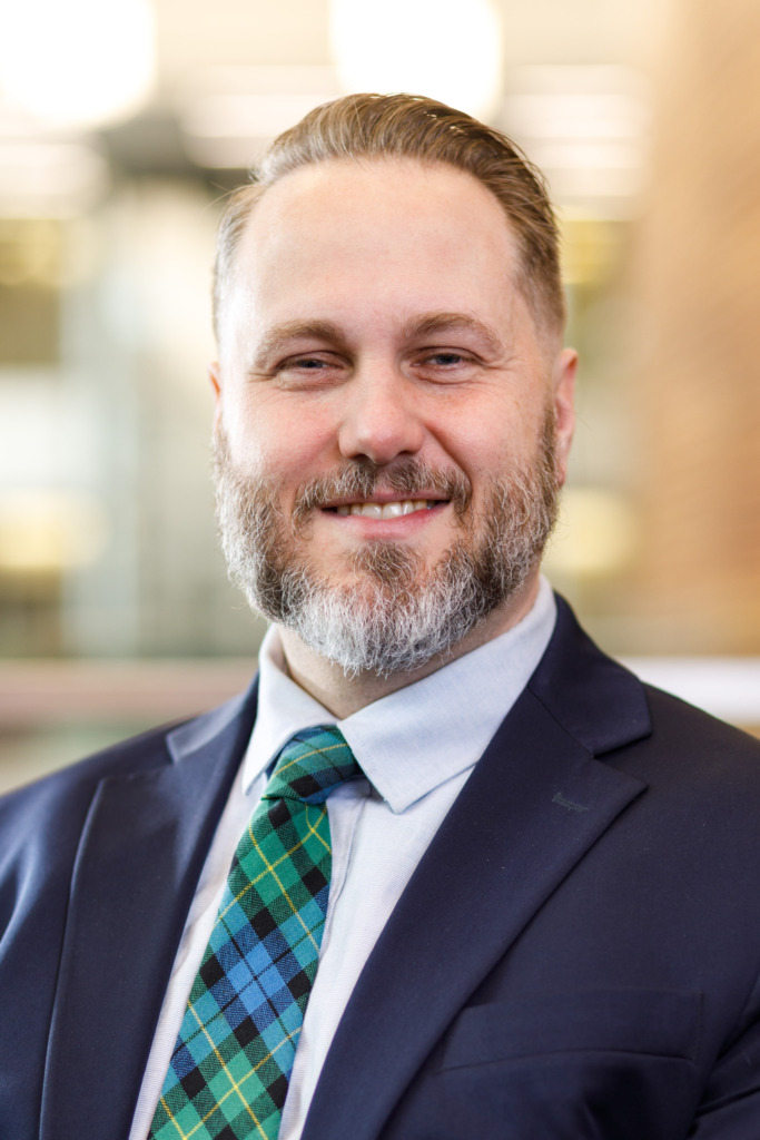A person with a beard and neatly styled hair smiles while wearing a dark suit, light blue shirt, and a green and blue plaid tie.