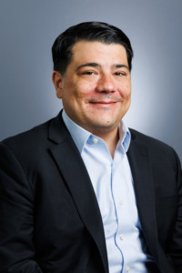 A professionally dressed individual with dark hair and a light blue shirt smiles warmly against a soft gray background.