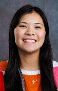 A person with long dark hair smiles warmly while wearing a colorful striped sweater and a small cross necklace.
