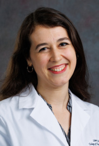A smiling individual in a white lab coat with embroidered text on the left chest, standing against a dark textured background.