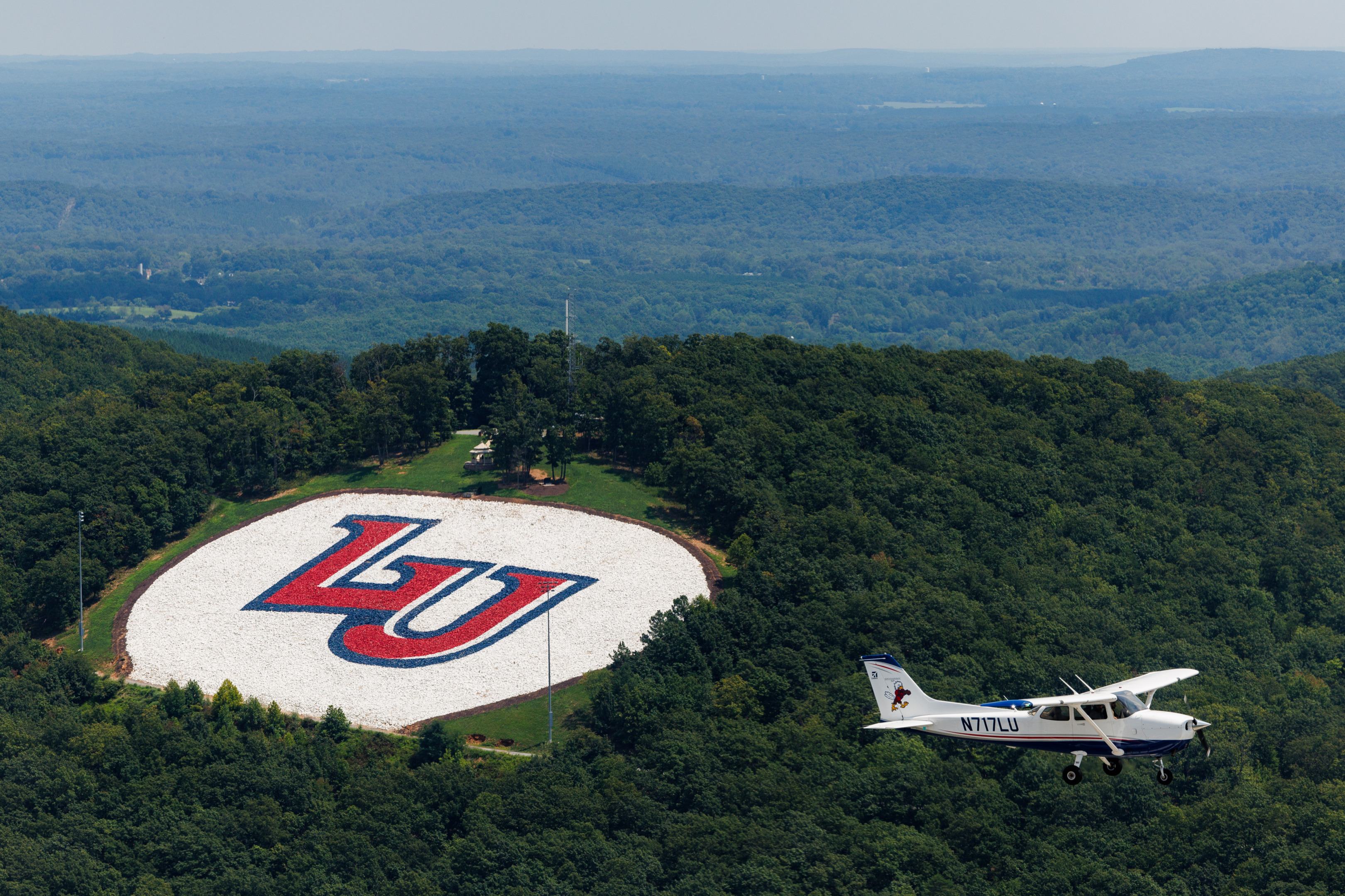 A small airplane flies over a large circular logo featuring the letters "LU" in red and blue, set within a white field surrounded by dense forest.