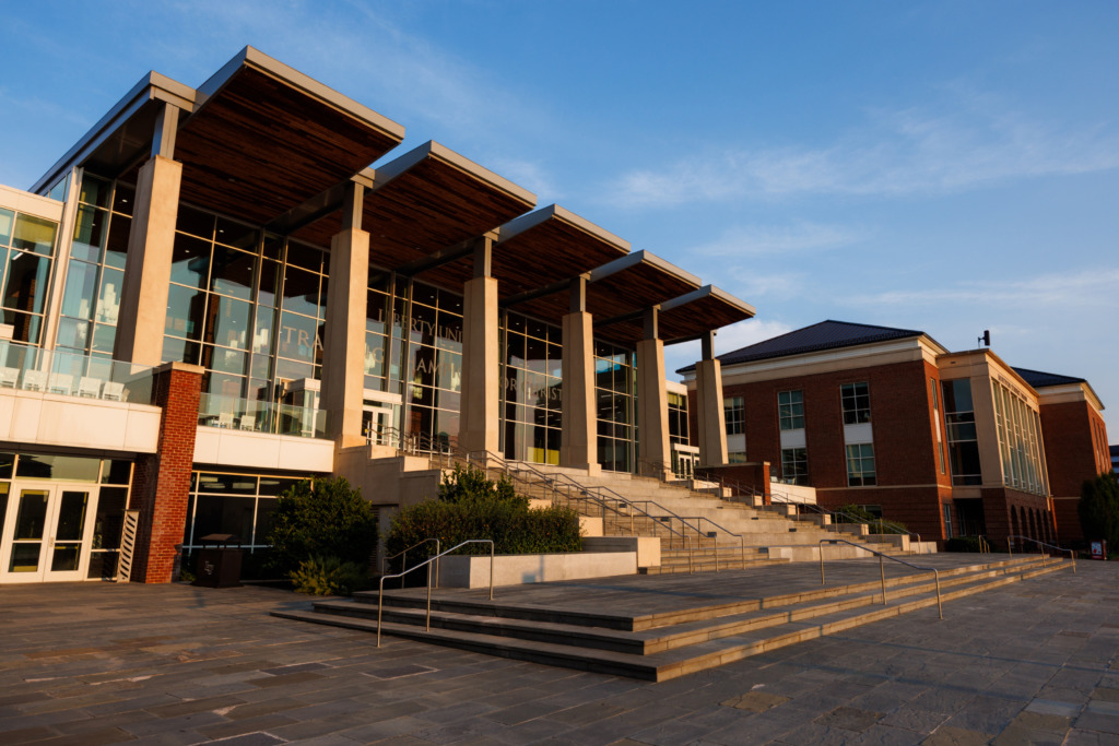 A modern building with large glass windows and wooden overhangs stands under a clear blue sky, featuring wide stone steps leading to its entrance.