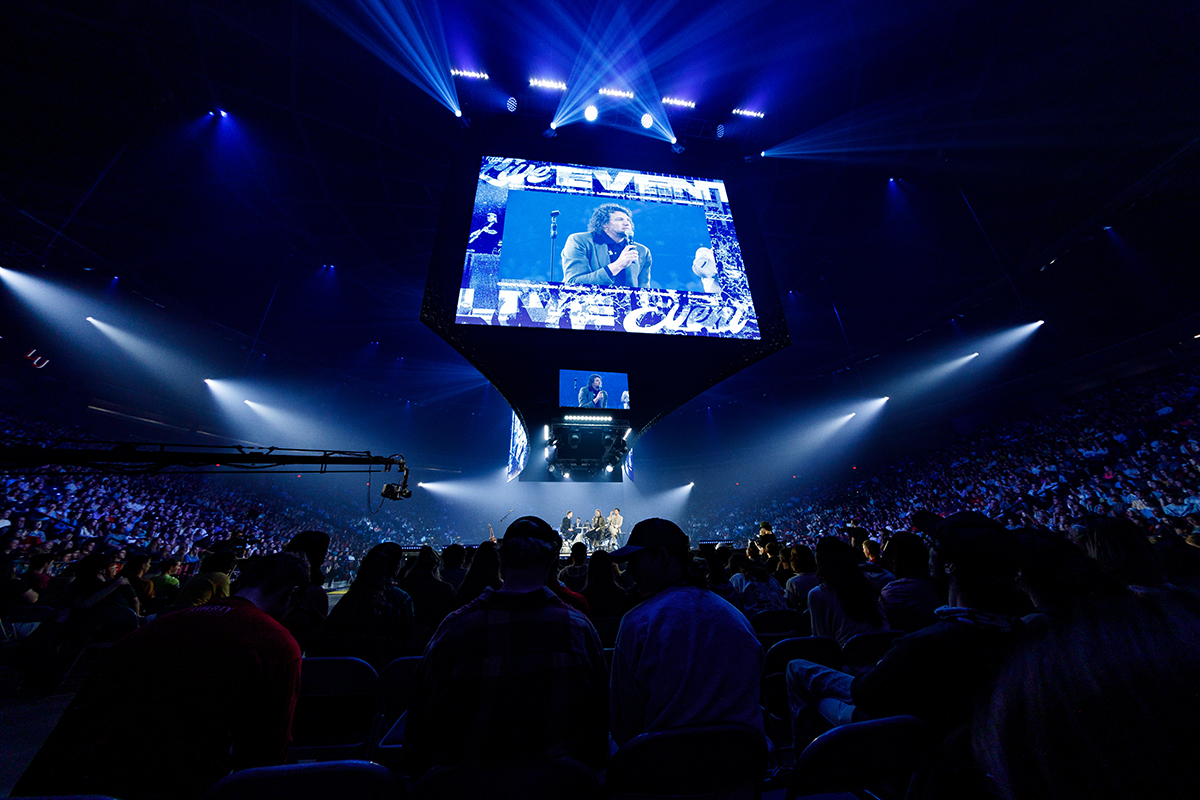 A large crowd watches a live performance on a brightly lit stage under blue spotlights, with a massive screen displaying the event.