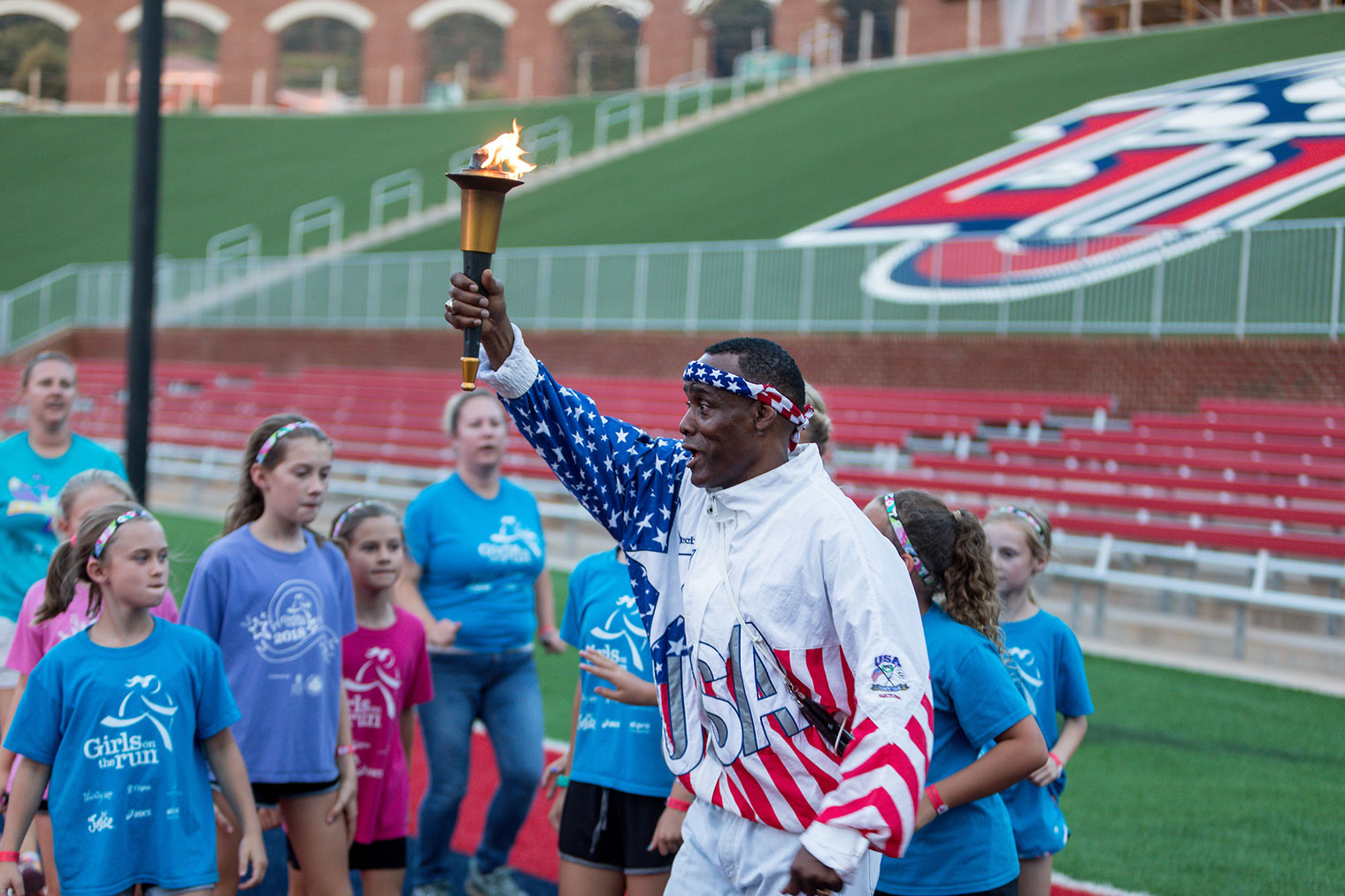 Anthony 'Buddy' Lee, who competed in the 1992 Summer Olympics in Barcelona and was a torch bearer for the 1996 Summer Games in Atlanta, receives the torch relayed into Williams Stadium by Girls On The Run on Friday night. 