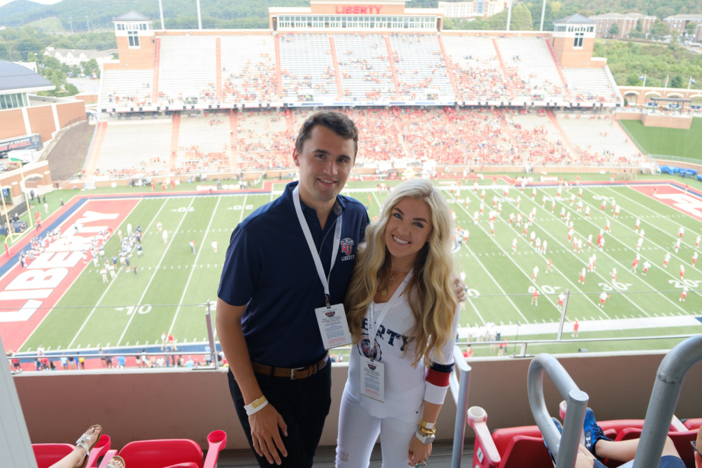 Two people pose together in the stands at a football stadium, with the field and spectators visible behind them.
