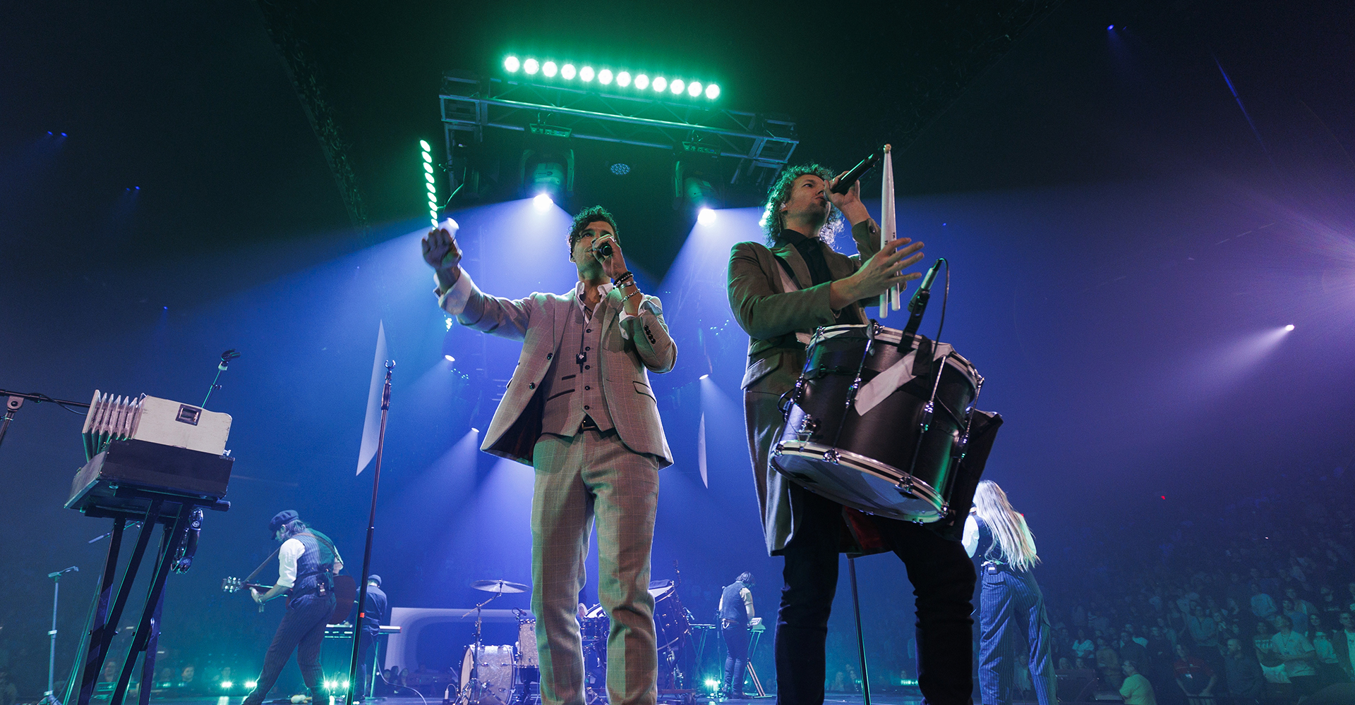 Two performers on a brightly lit stage, one singing into a microphone and the other playing a drum, with musicians and equipment visible in the background.