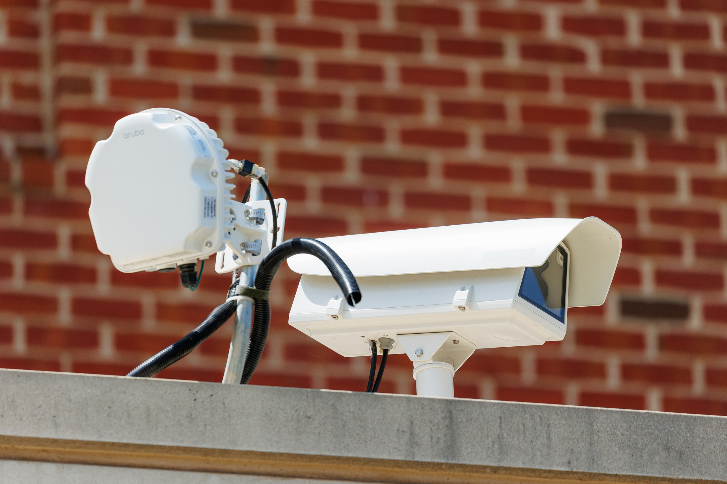 A white security camera and a wireless access point are mounted on a concrete ledge against a red brick wall.