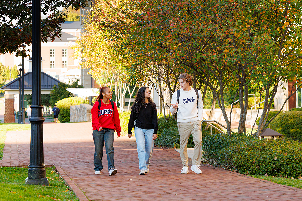 Three students walk along a brick pathway on a university campus, surrounded by autumn-colored trees and manicured greenery.