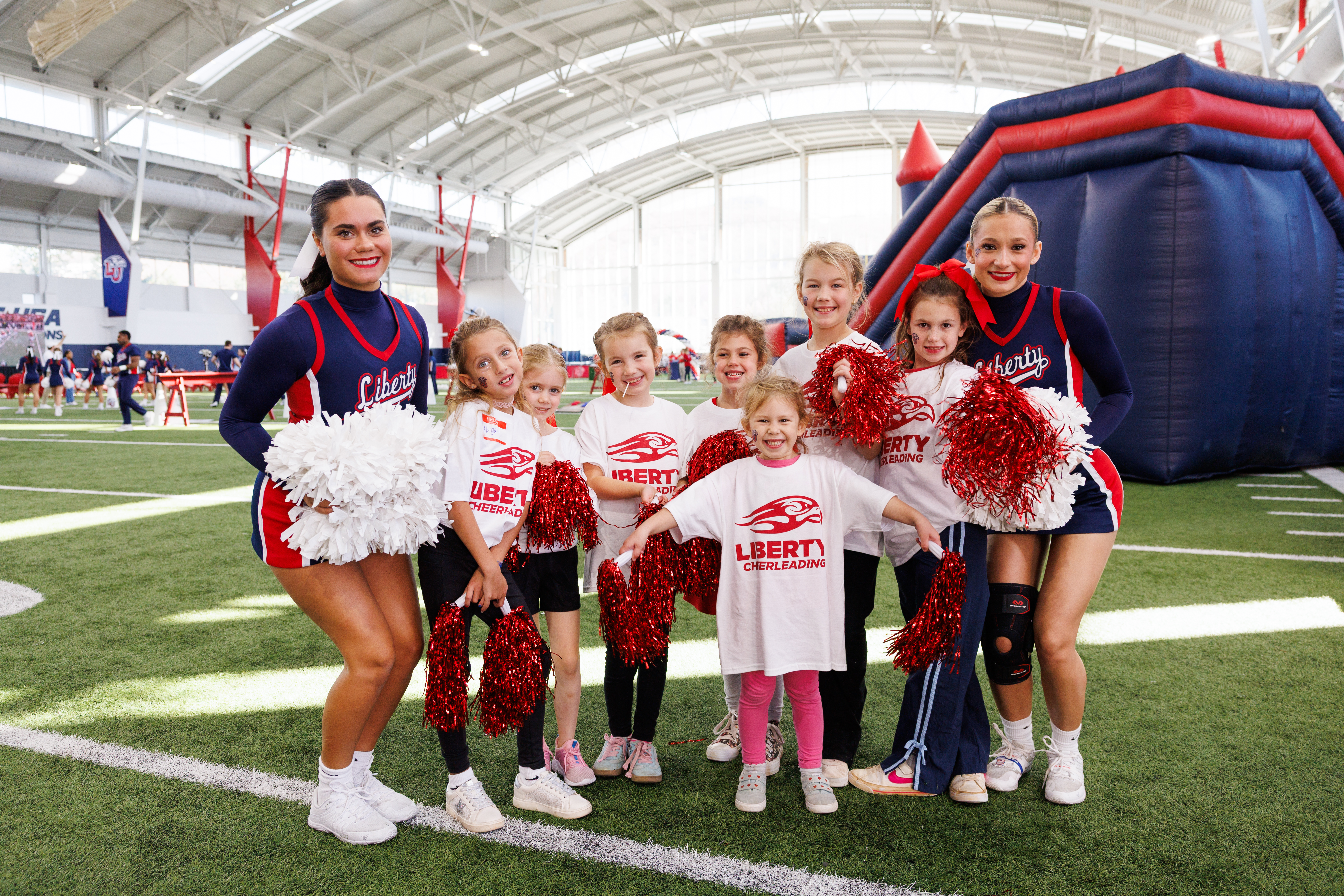 A group of cheerleaders and young children pose together on an indoor sports field, holding pom-poms and wearing matching team apparel.