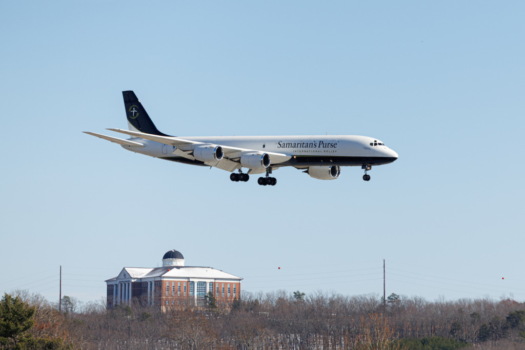 A Samaritan's Purse aircraft is in flight with landing gear down, approaching an airport against a clear blue sky, with a building and trees visible below.