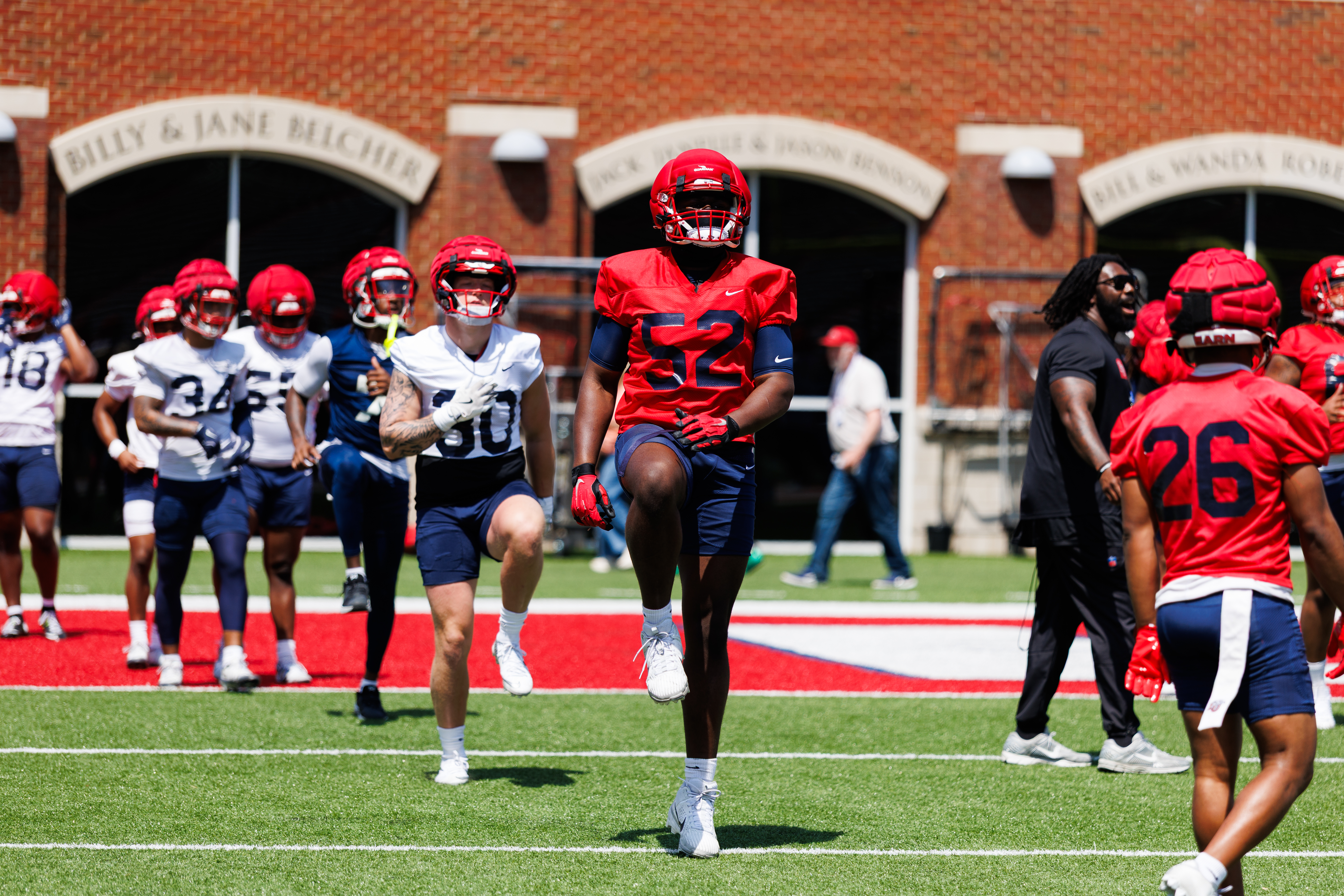 Football players in red and white uniforms run on a field during practice, with a brick building featuring arched entrances in the background.