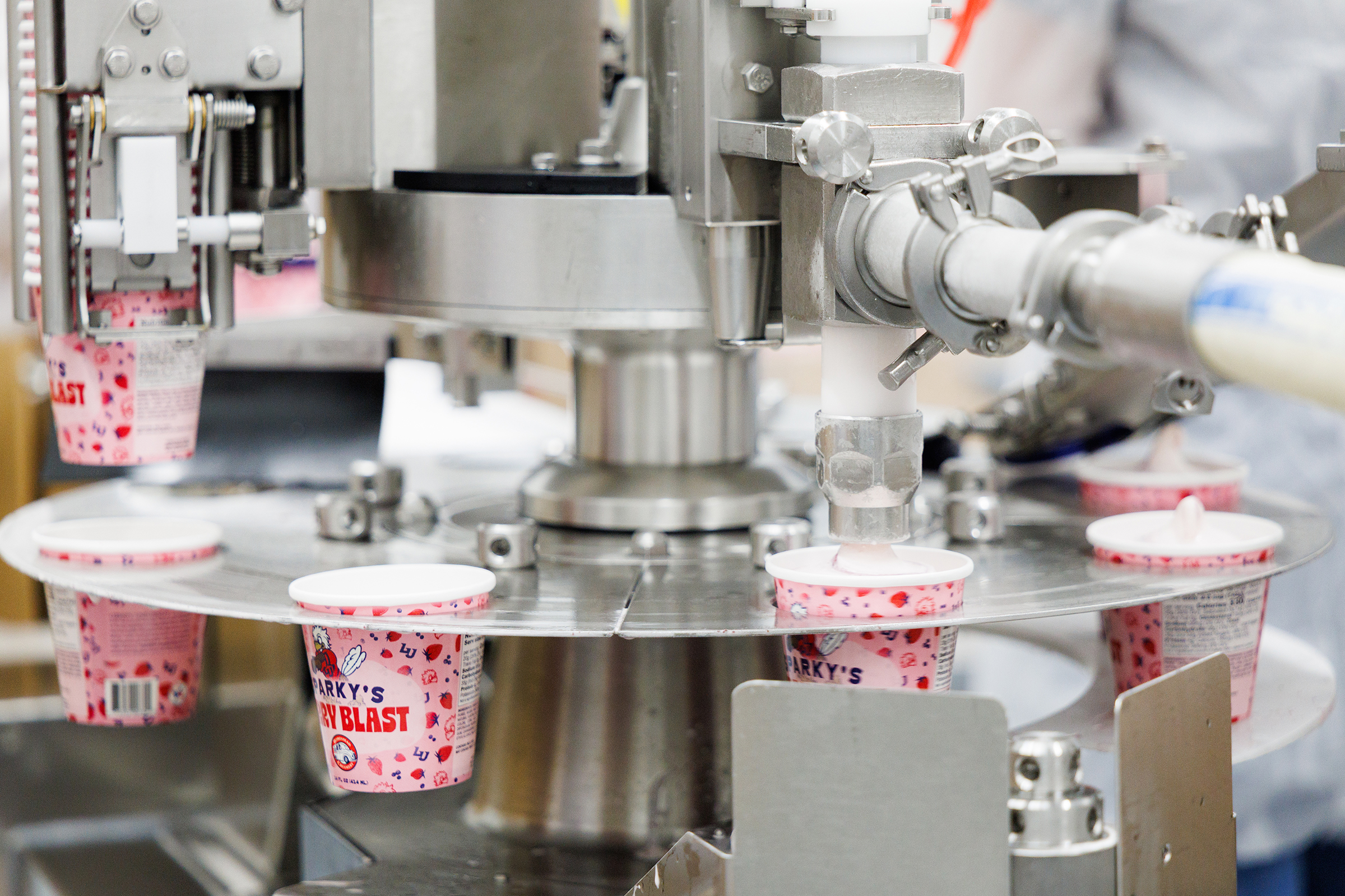 A robotic machine fills strawberry-flavored yogurt cups with a creamy substance in a factory setting.