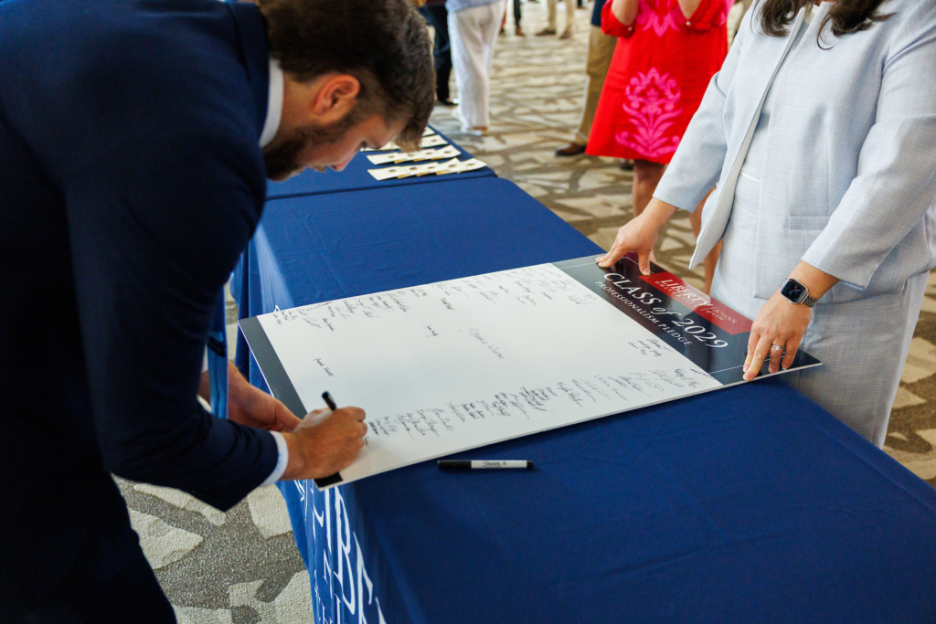 A person in a suit signs a large document at a table covered with a blue cloth, while another individual holds the document steady.