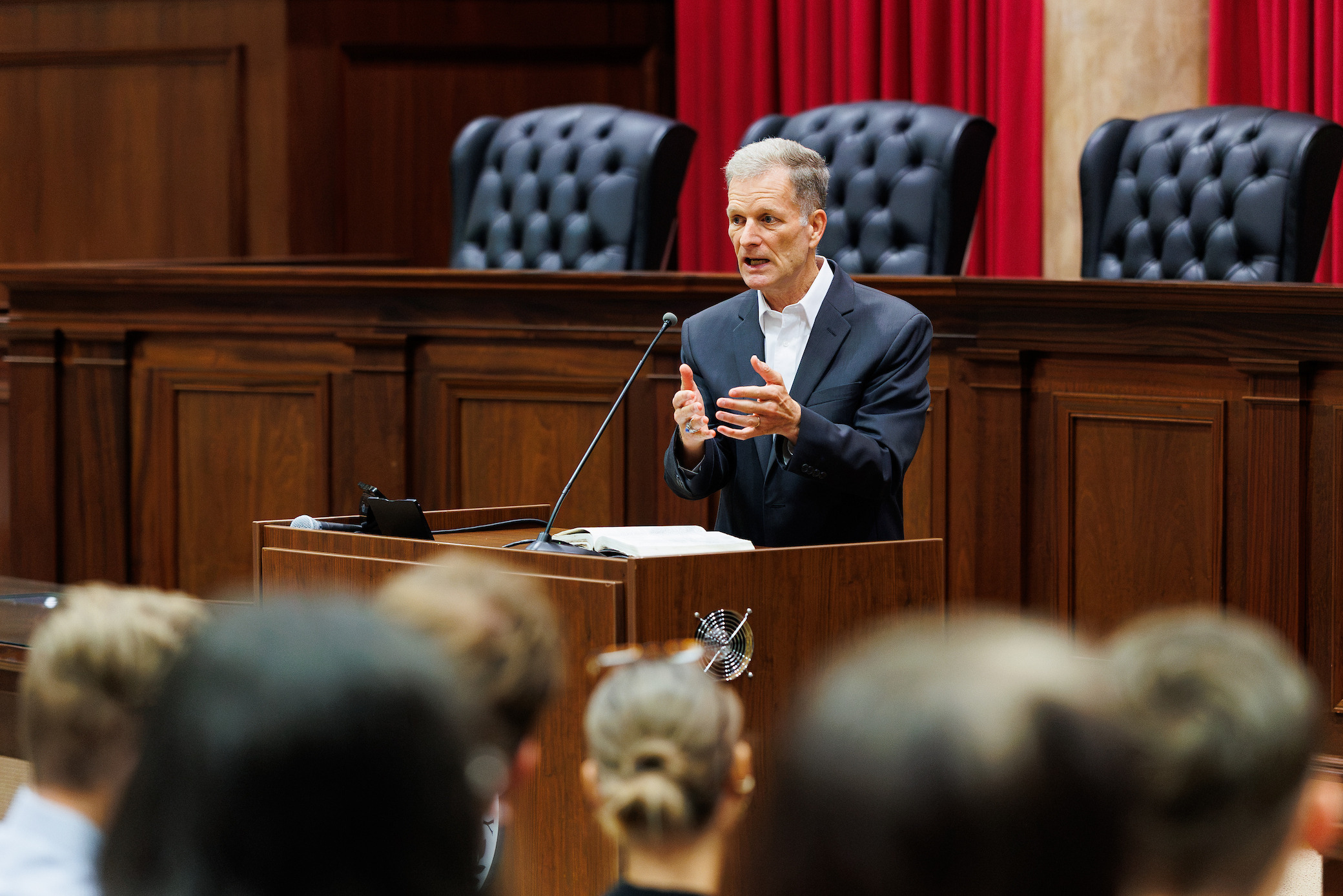 A person in a suit stands at a wooden podium, speaking to an audience in a formal room with red curtains and upholstered chairs.