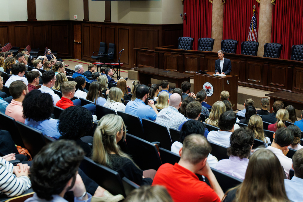 A speaker addresses an audience in a formal lecture hall with red curtains and a large screen displaying event details.