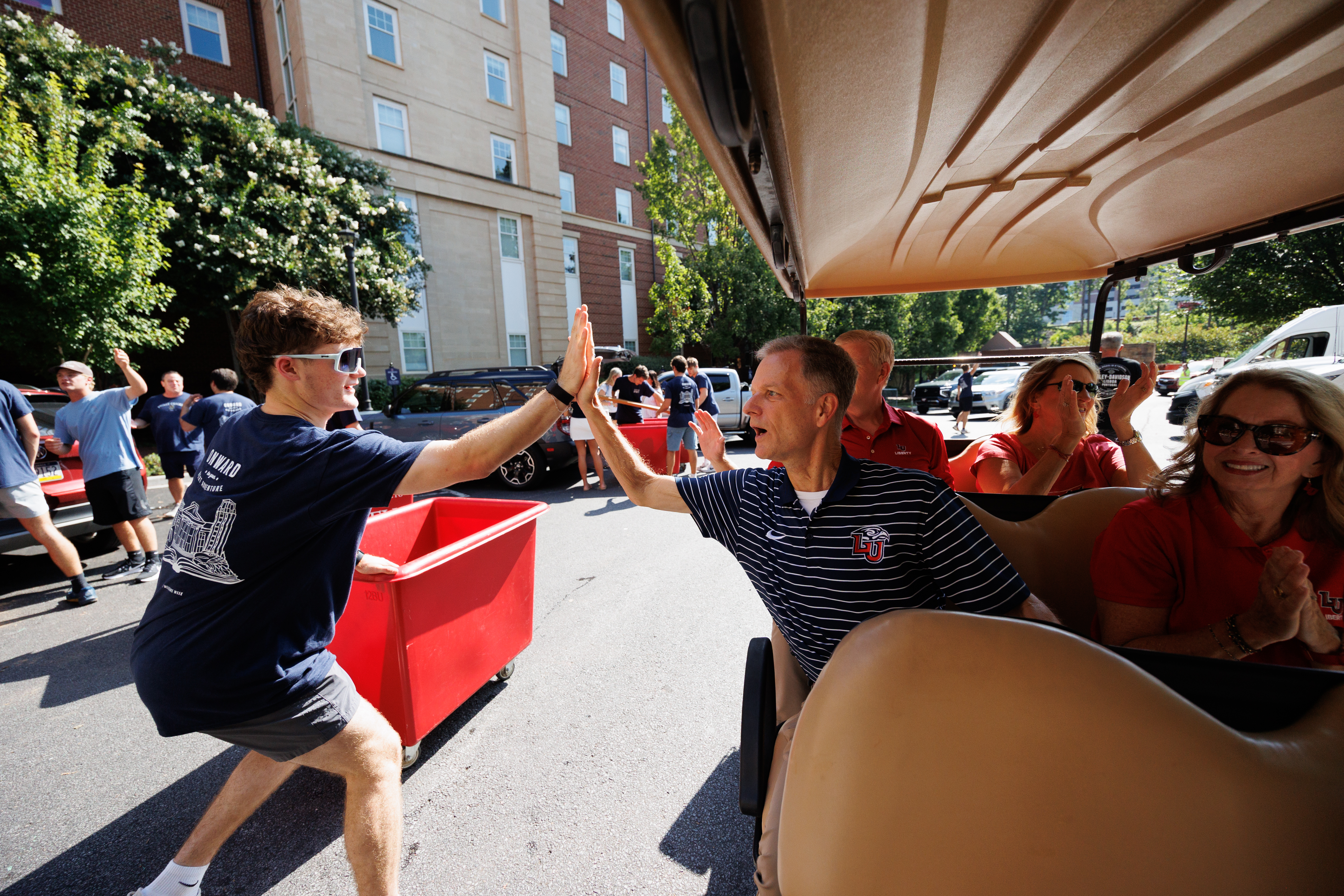 A young person in a navy shirt gives a high-five to an adult in a striped polo shirt while seated in a golf cart, with others watching and cheering nearby.