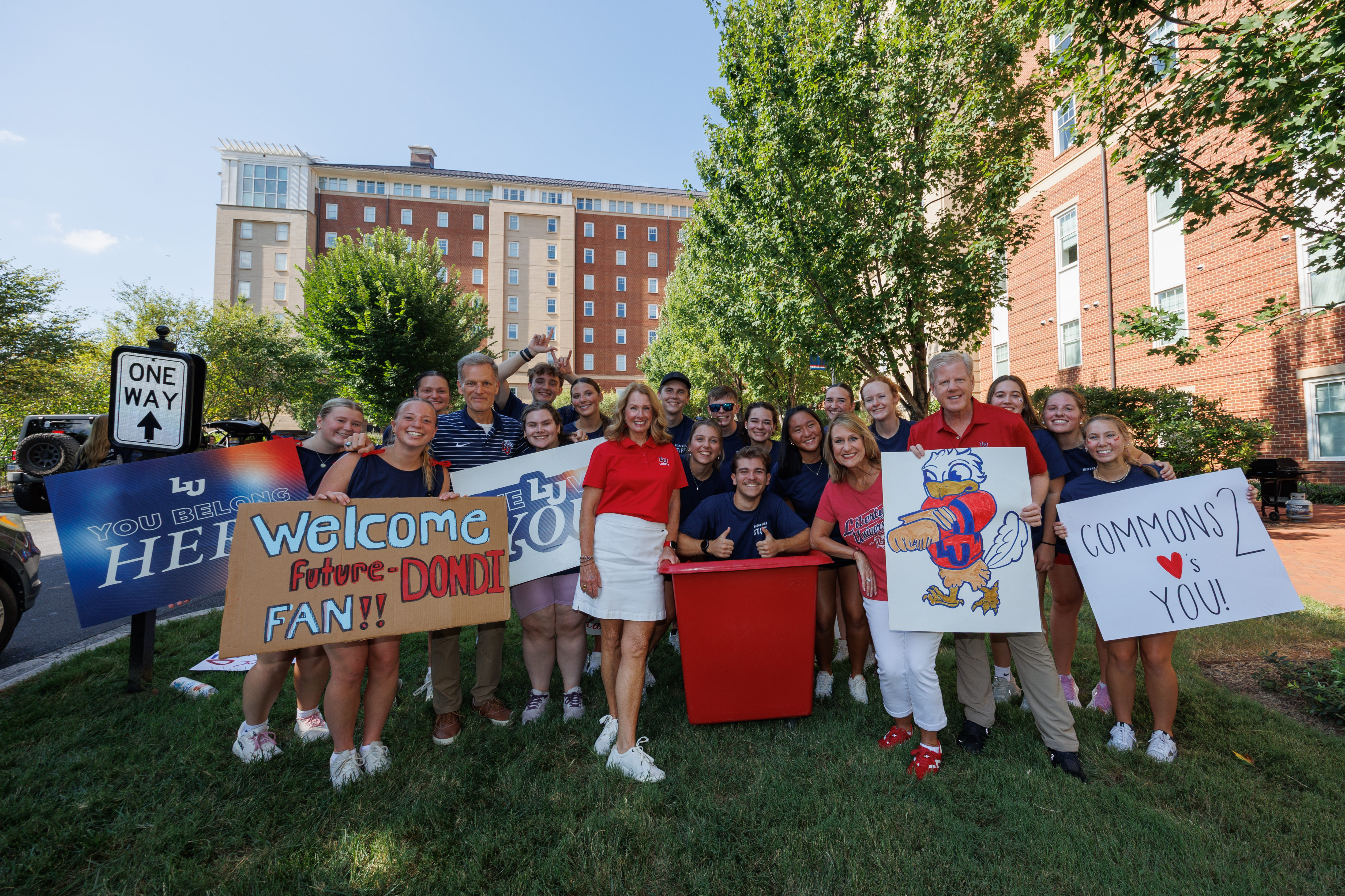 A group of people, including a man in a red polo shirt and a woman in a red top, stand on grass holding signs that welcome a future fan and promote the university's commons, with a large brick building and trees in the background.