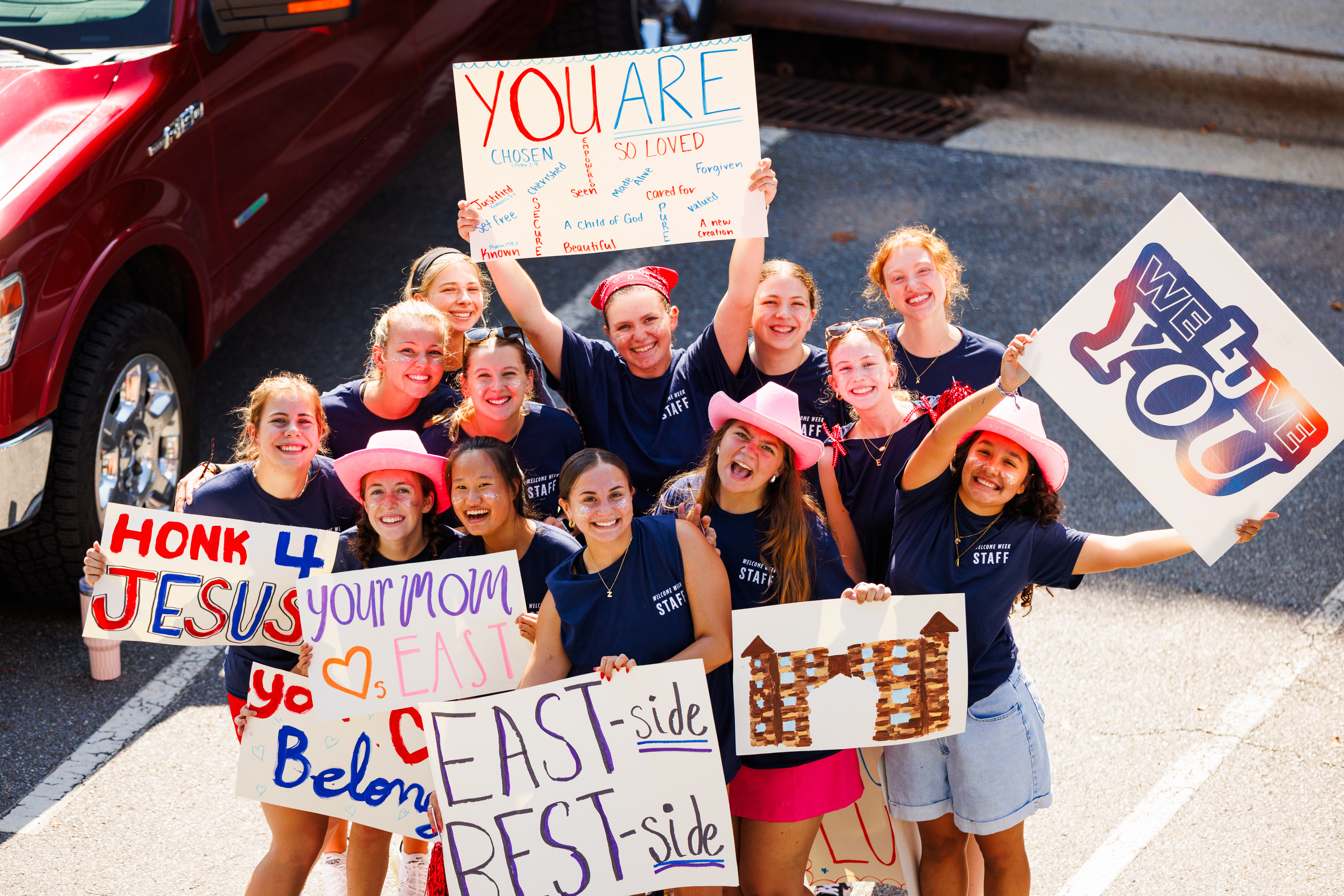 A group of smiling individuals in matching shirts and pink hats hold colorful signs with uplifting messages, standing outdoors near a red vehicle.