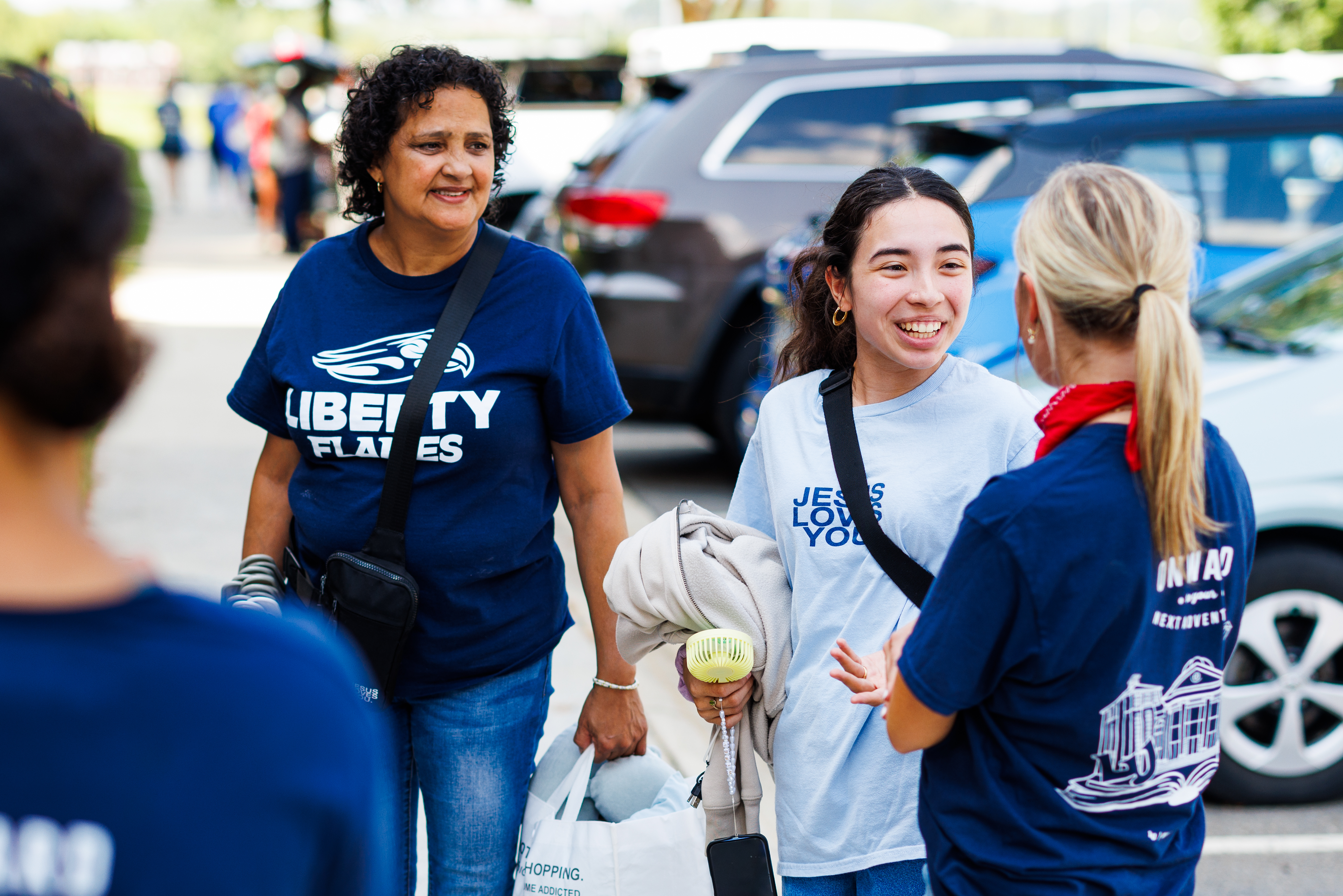 Three individuals in matching blue shirts converse outdoors near parked cars, with one holding a shopping bag and another carrying a small fan.