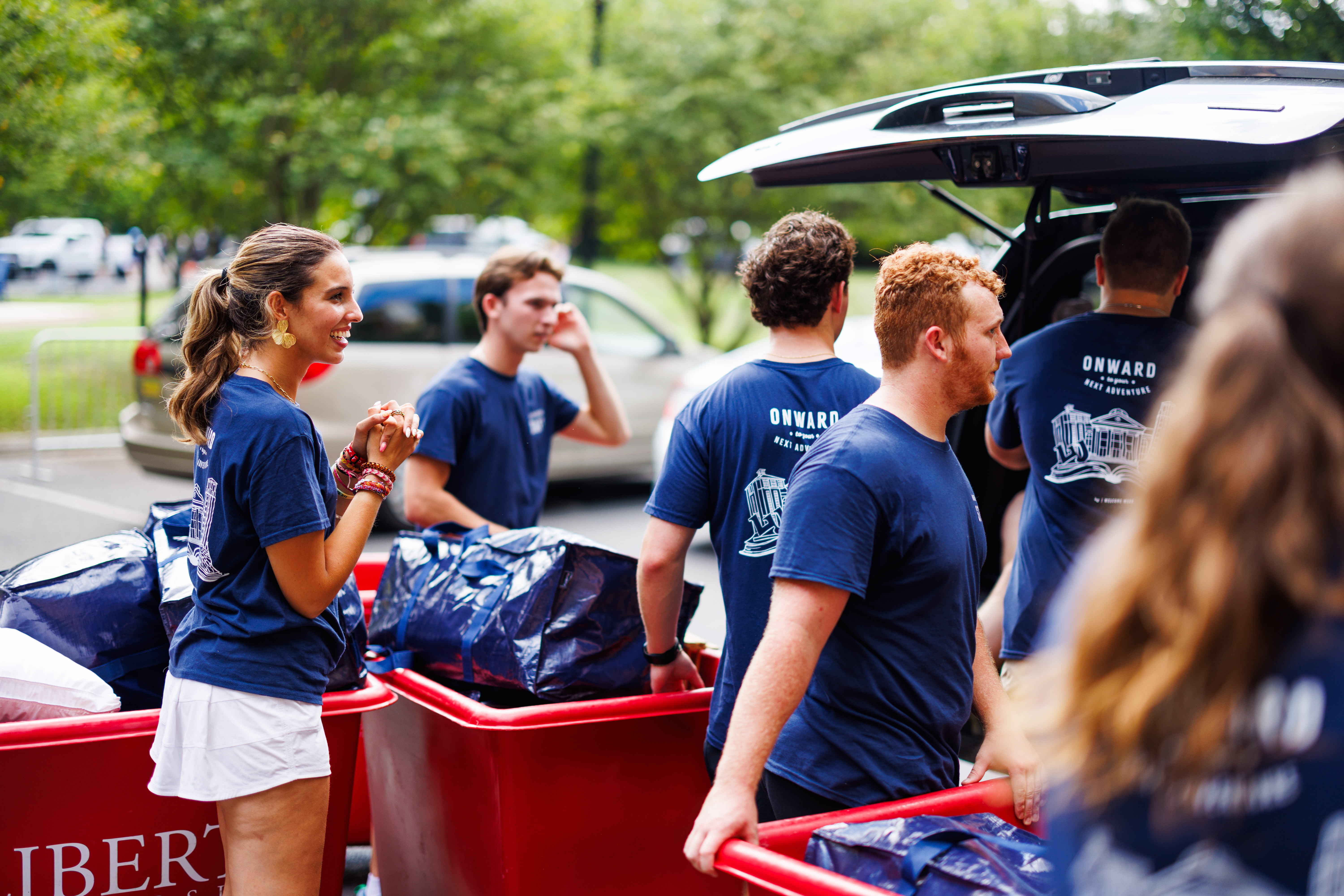 A group of people in matching blue shirts load large blue bags into red bins near an open car trunk on a sunny day.