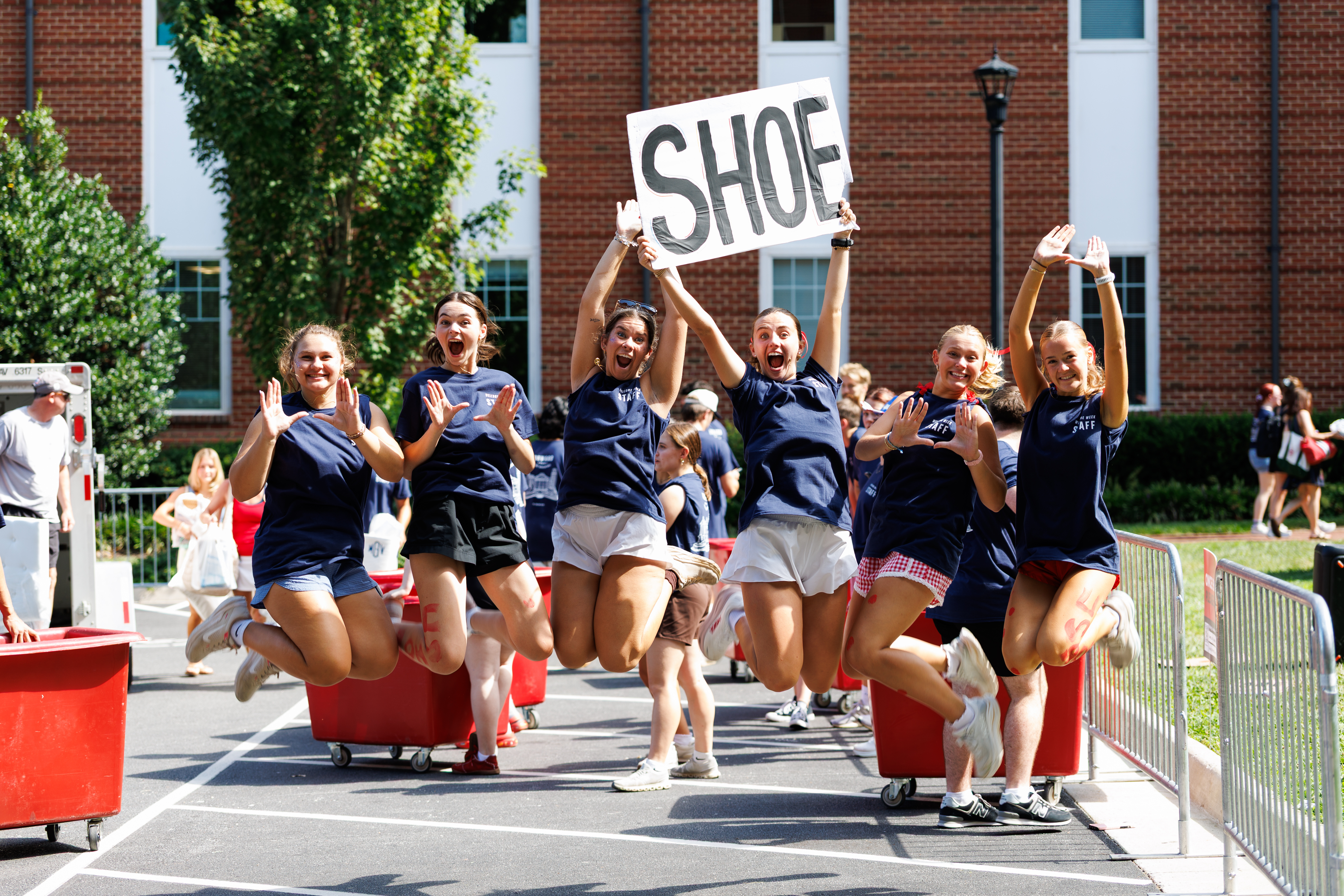 A group of young people jump joyfully in unison, holding a sign that reads "SHOE," while standing beside red carts on a paved area near a brick building.