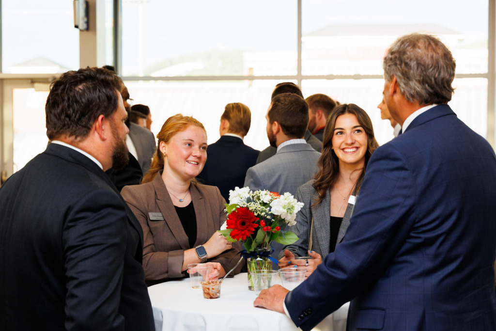 Several individuals in business attire engage in conversation around a table adorned with a floral centerpiece at a professional gathering.