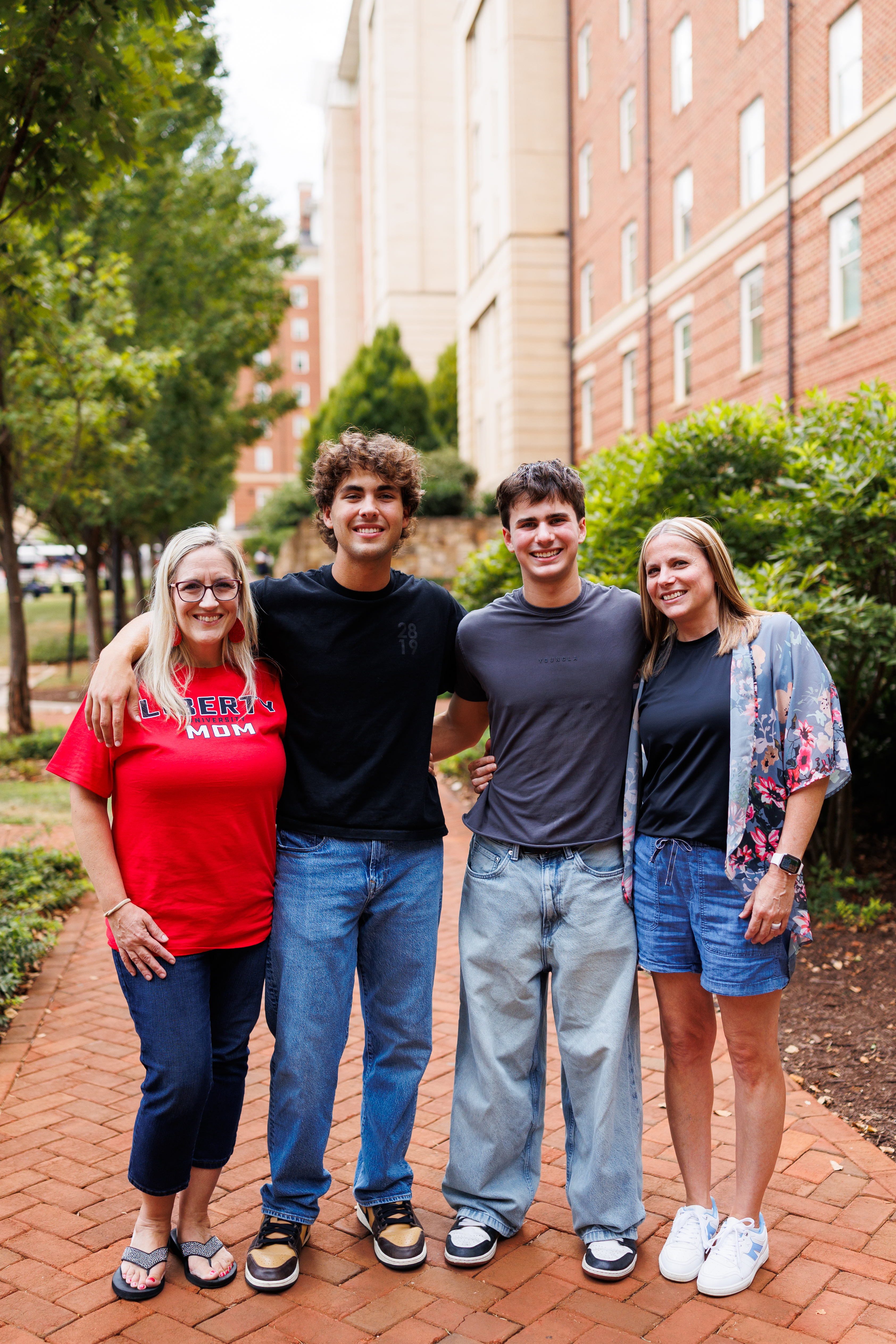 A family of four smiles together on a brick pathway, with two adults and two young people standing close, surrounded by trees and a brick building in the background.