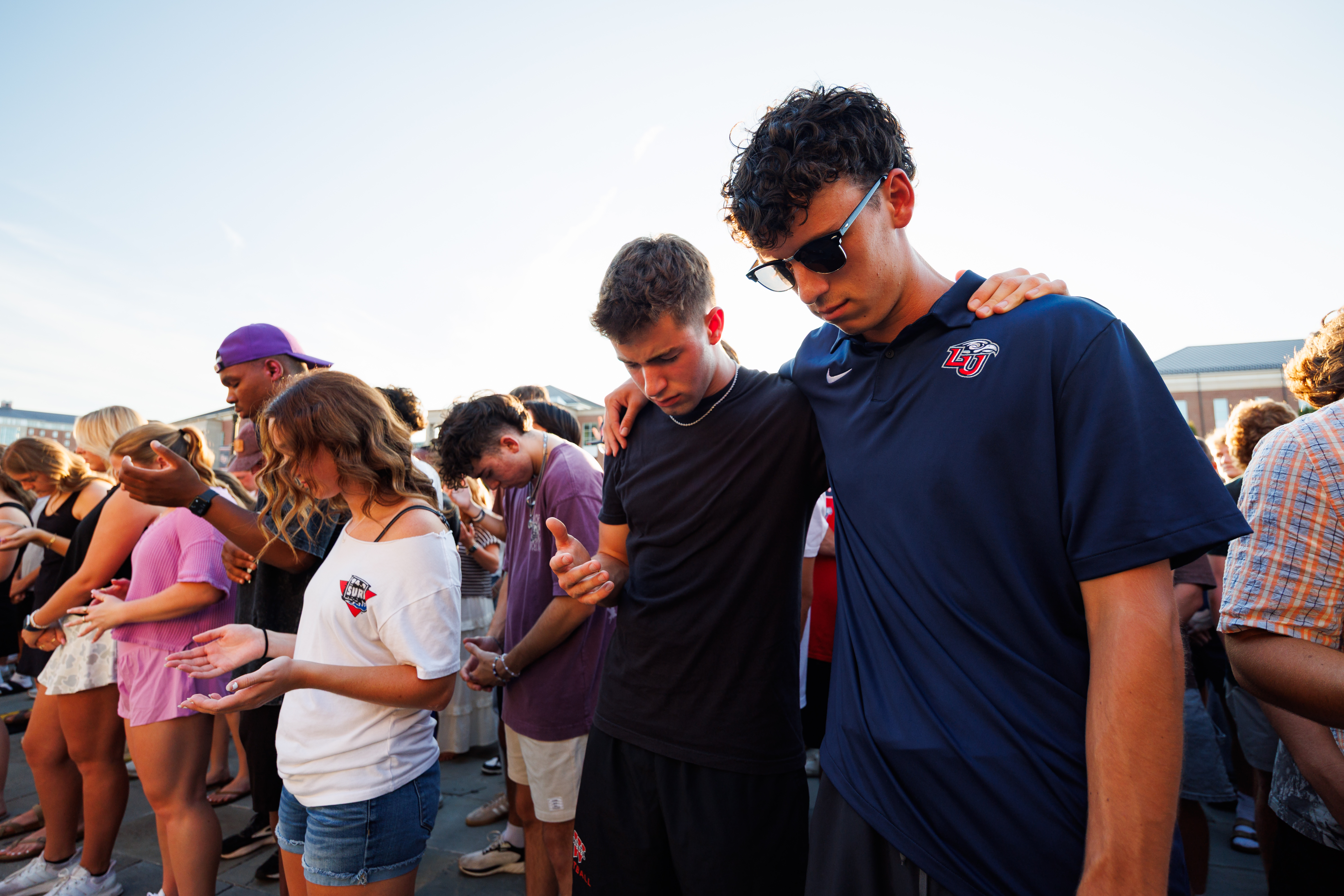A group of young people stand in a line outdoors, heads bowed and hands clasped, appearing to be in prayer or reflection.