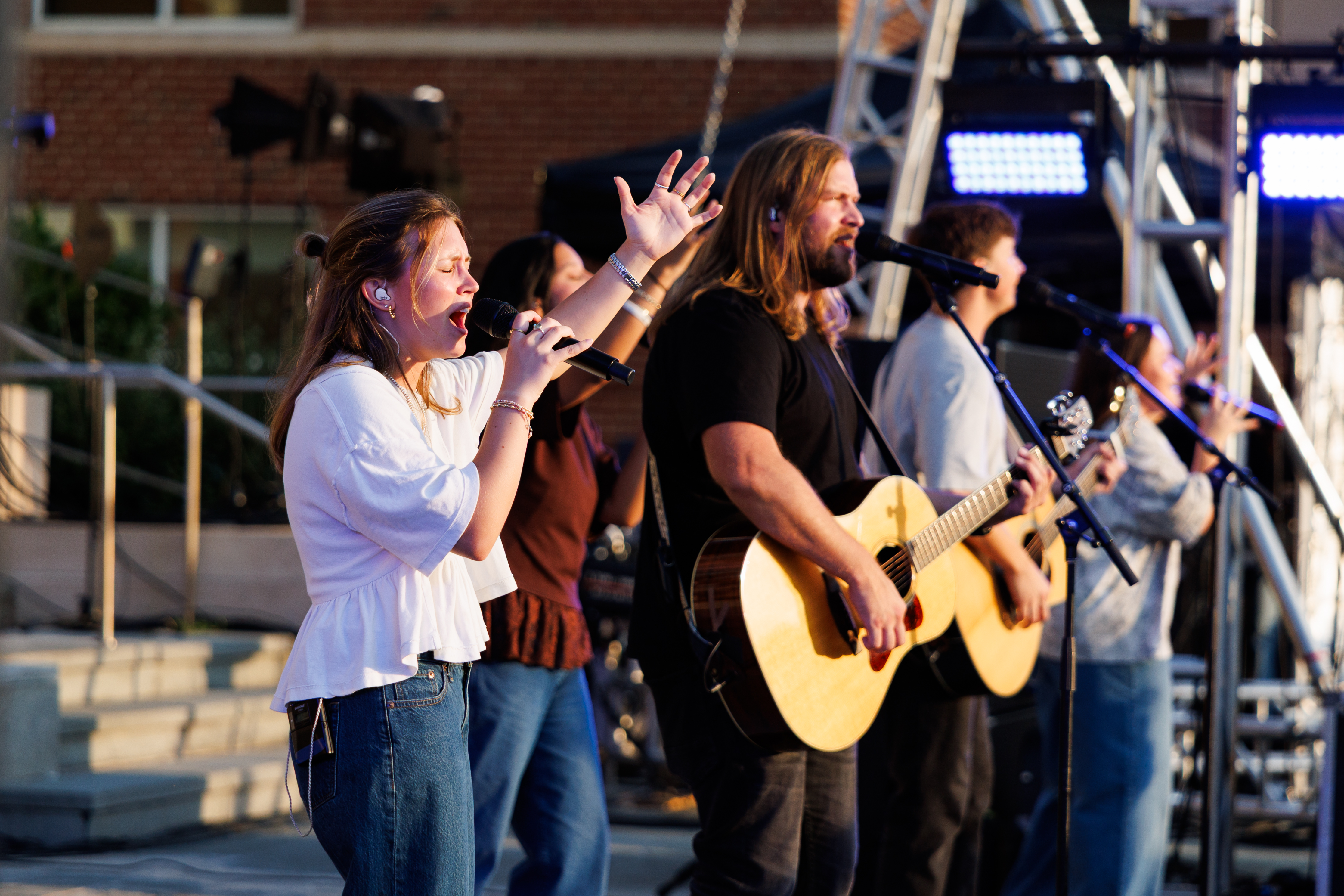 A group of musicians performs on an outdoor stage, with one singer holding a microphone and another playing an acoustic guitar.