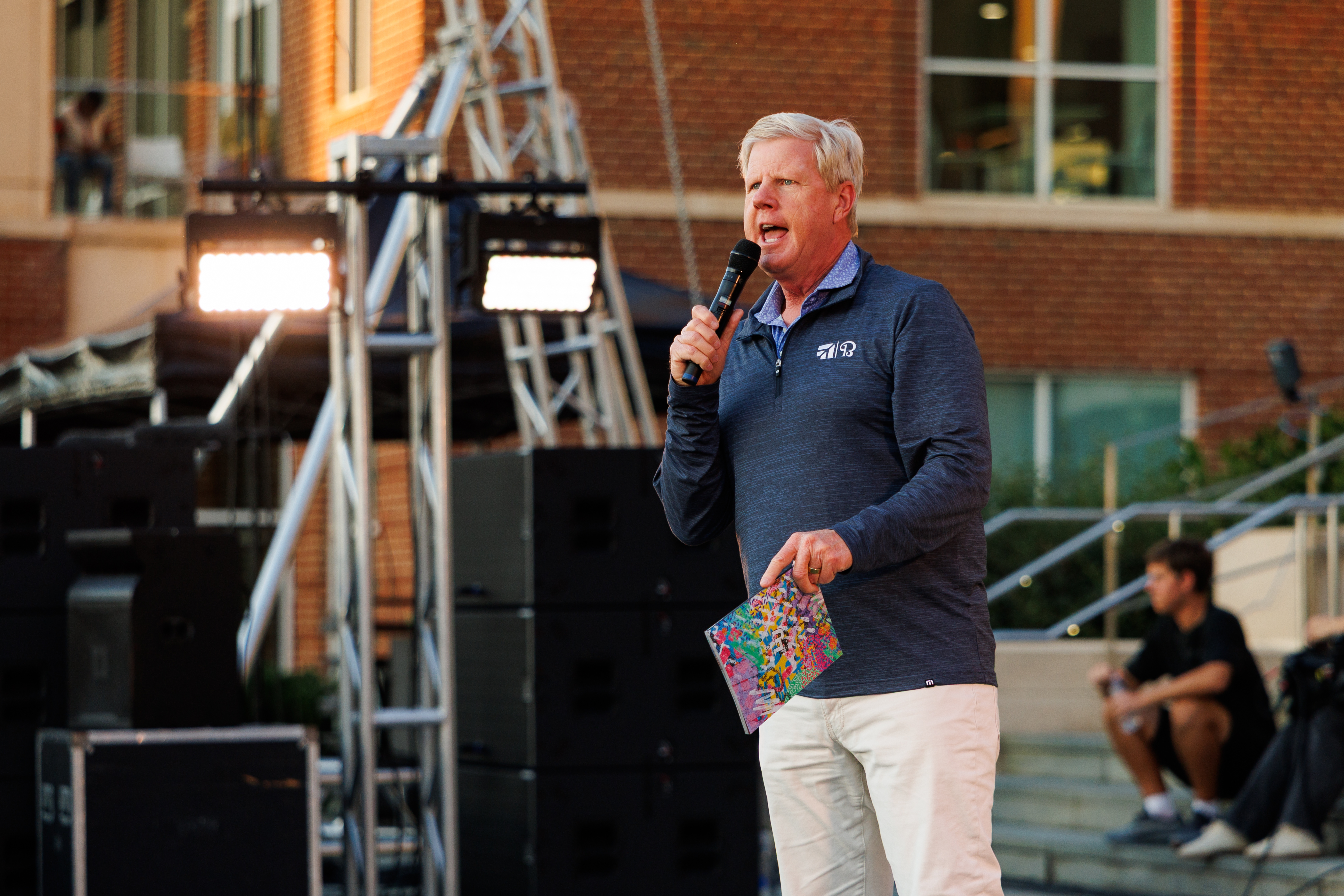 A person speaks into a microphone on a stage, holding a colorful booklet, with event lighting and sound equipment visible.