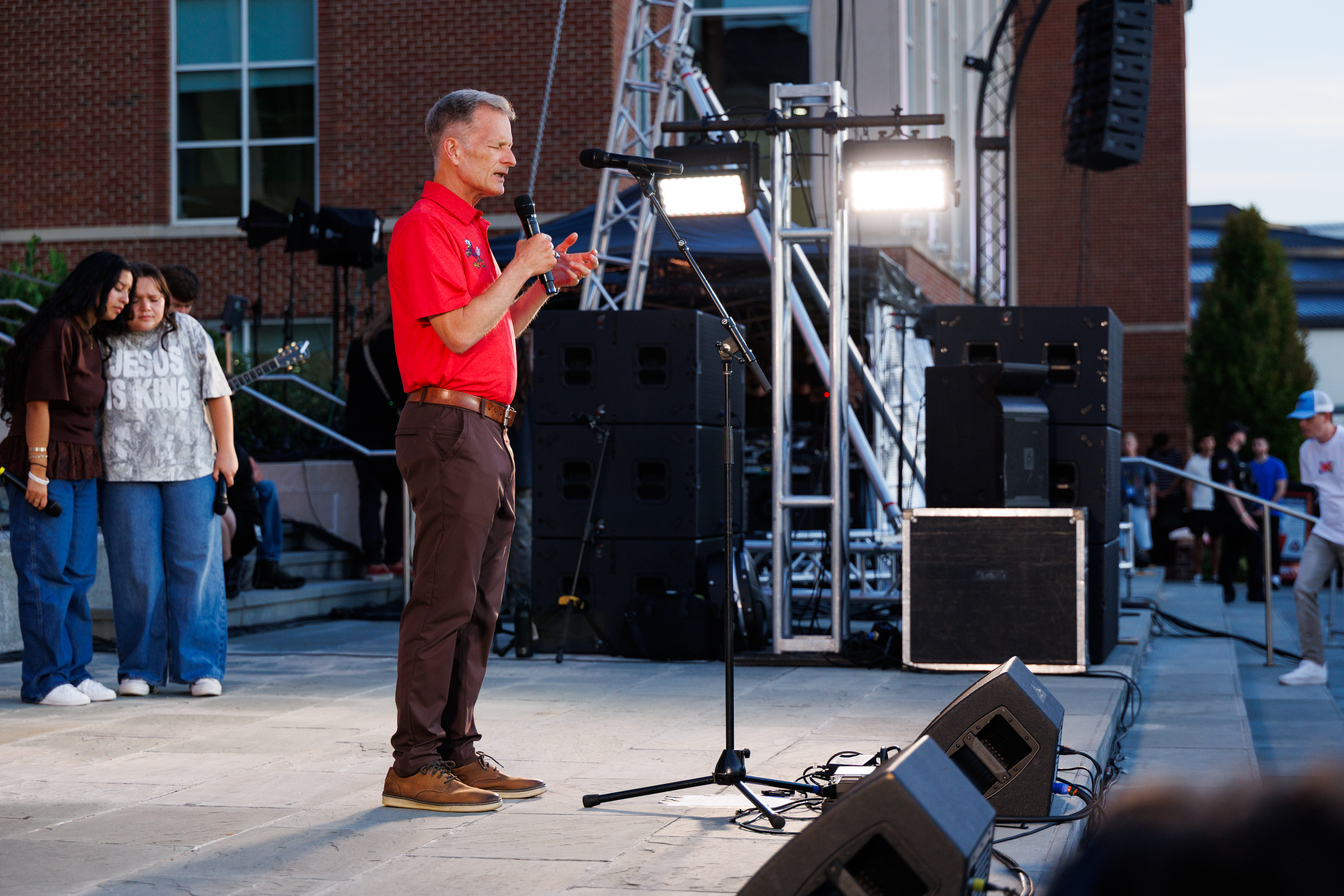 A person in a red shirt speaks into a microphone on a stage, with large speakers and lighting equipment around them, while others stand nearby.