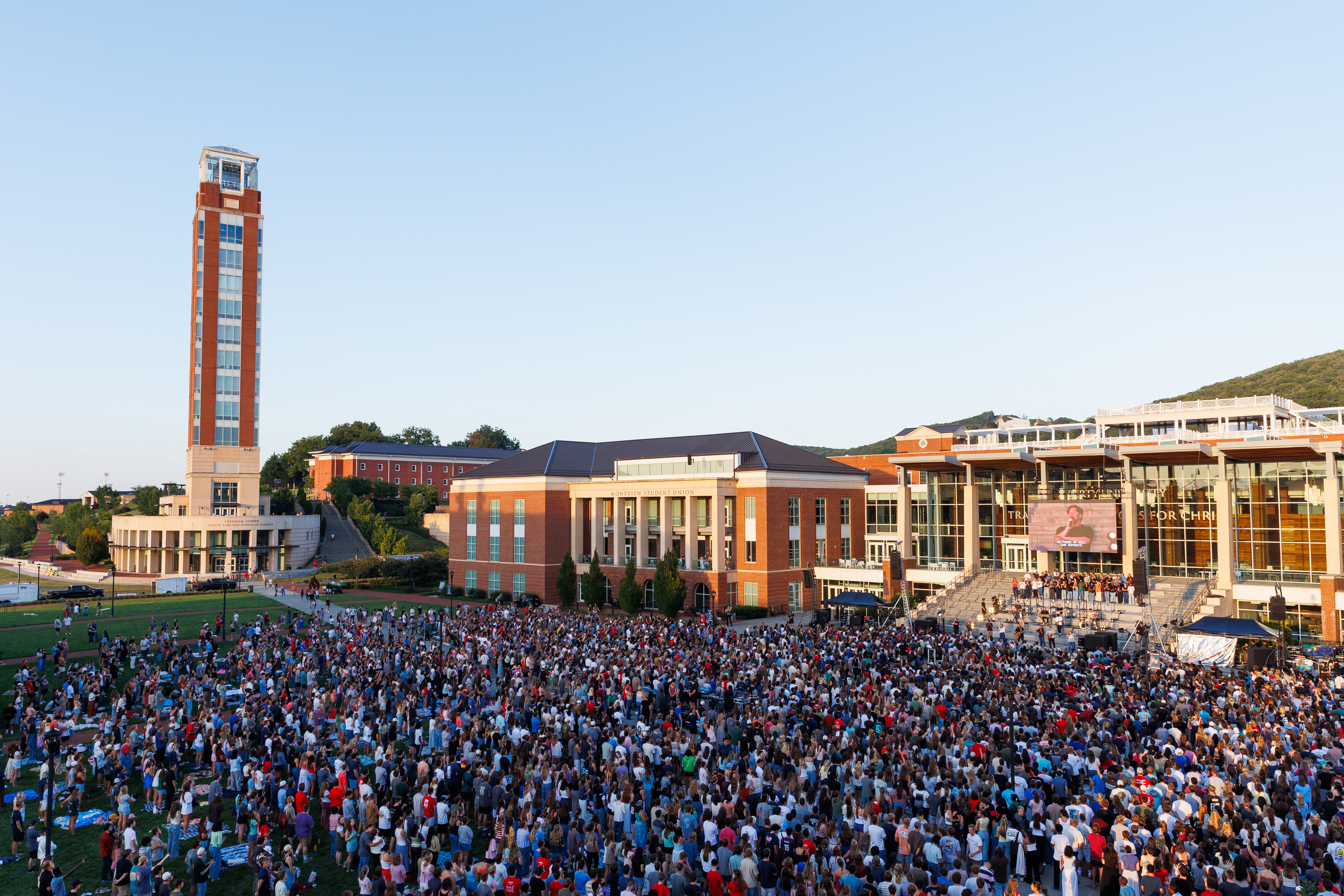 A large crowd gathers on a grassy field in front of a university campus with a tall tower and modern buildings under a clear sky.