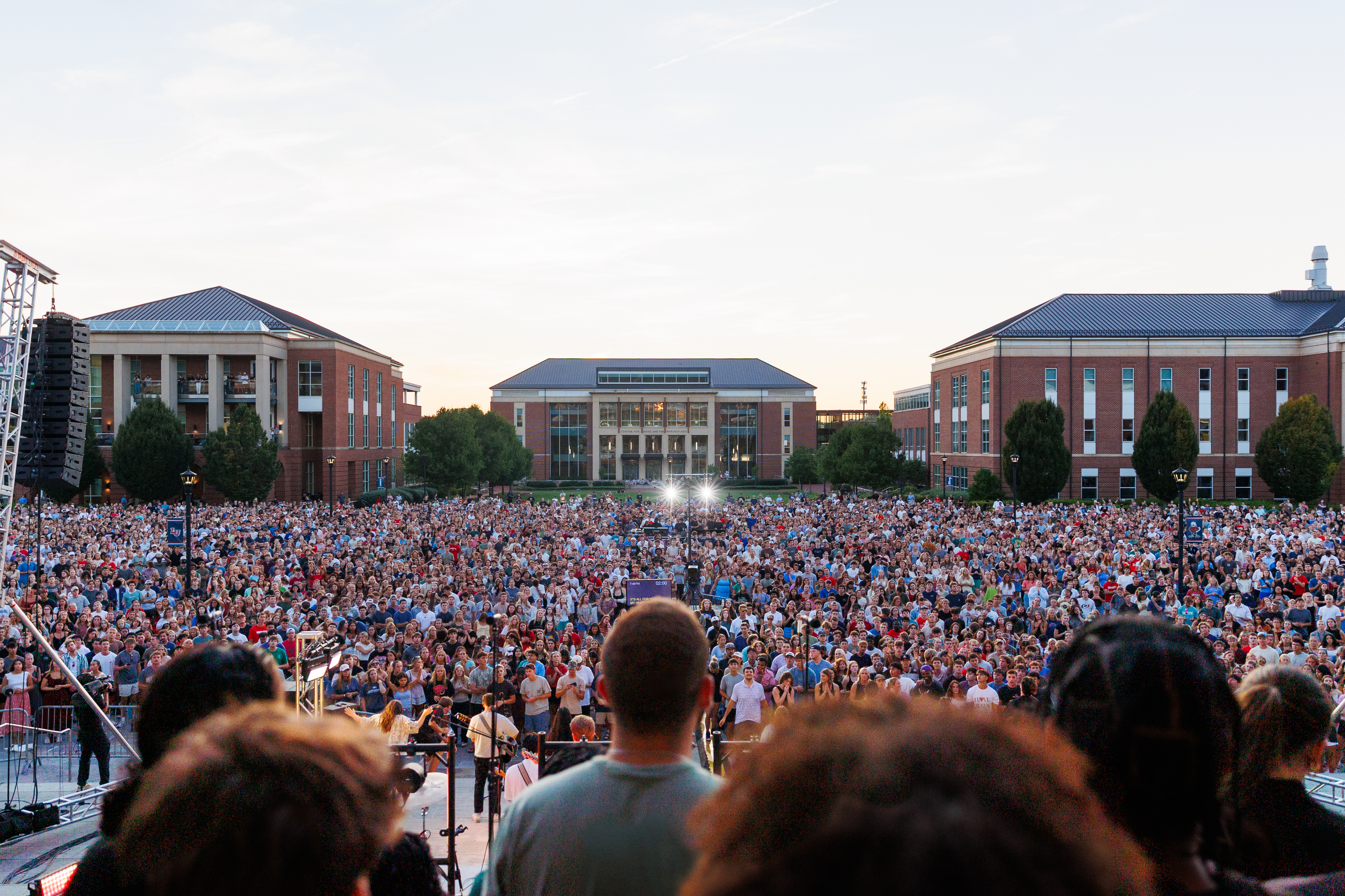 A large crowd gathers in an open square at a university campus for an outdoor concert, with buildings and stage lights visible in the background.